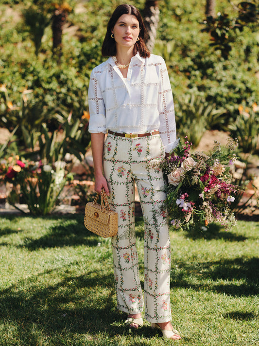 Model wearing J.McLaughlin Helen Linen Blend Pants in Floral Medley Frame paired with a white blouse, holding a woven handbag and a bouquet of flowers in a garden setting, showcasing effortlessly stylish, enduring designs with innovative fabrics for a life well-lived.