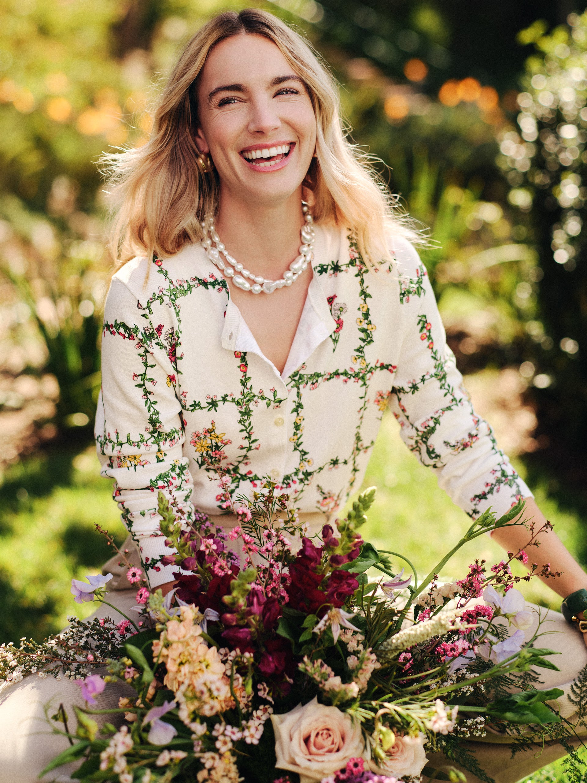 Smiling woman outdoors wearing the Whitney Cardigan in Floral Medley Frame by J.McLaughlin, surrounded by vibrant fresh flowers, effortlessly stylish with enduring designs.