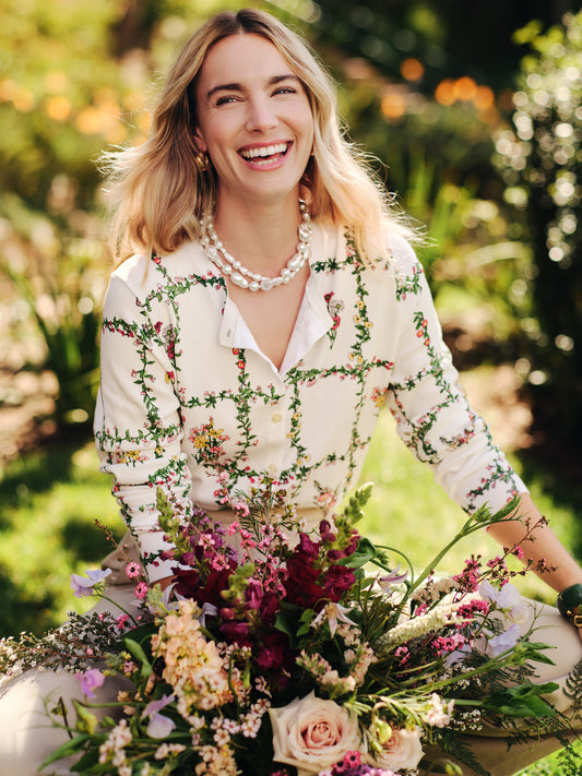 Smiling woman outdoors wearing the Whitney Cardigan in Floral Medley Frame by J.McLaughlin, surrounded by vibrant fresh flowers, effortlessly stylish with enduring designs.