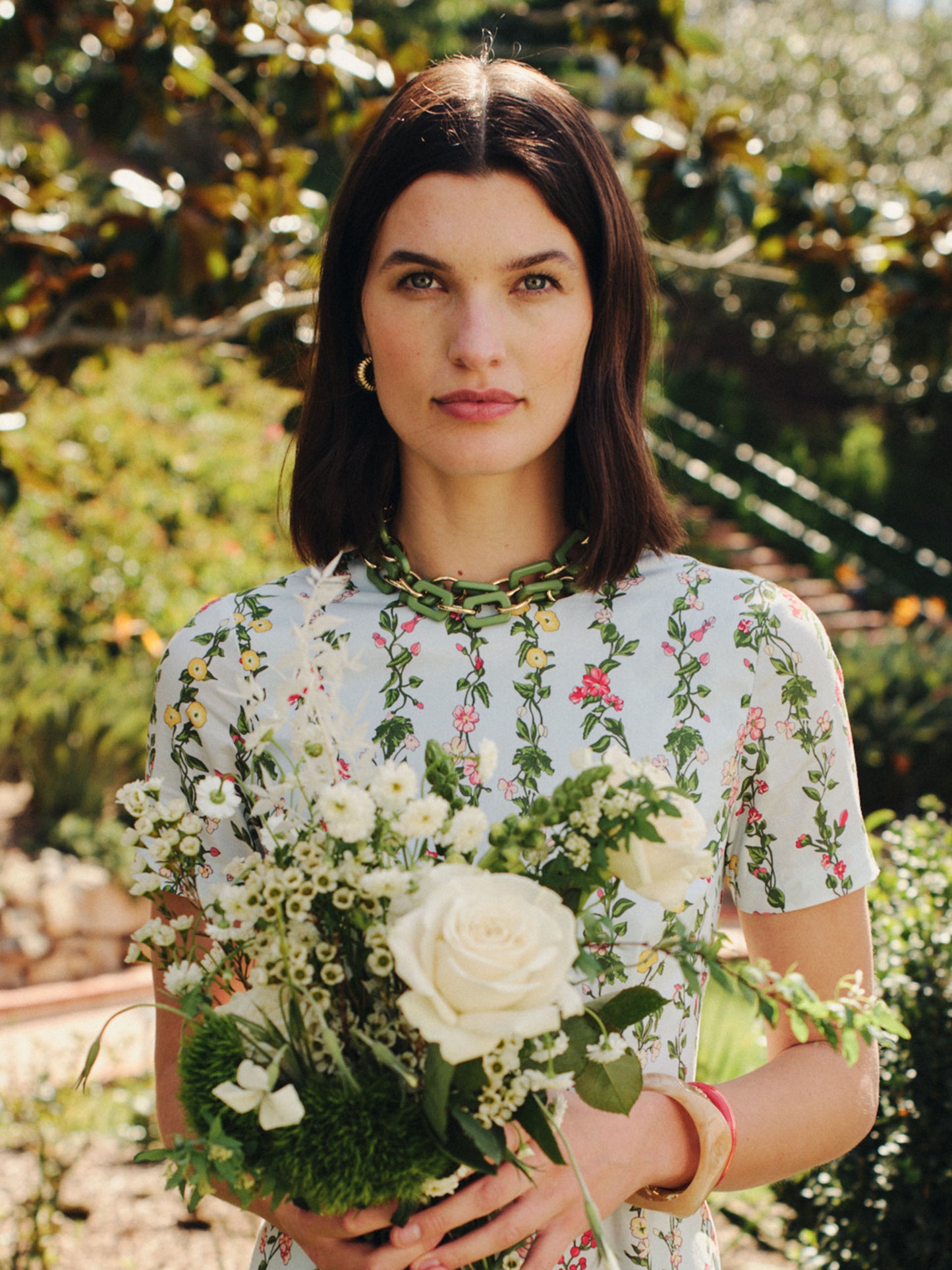 Woman wearing a floral dress and a Chain Link Necklace by J.McLaughlin, holding a bouquet of white roses and greenery in a sunlit garden, embodying effortlessly stylish, enduring designs.