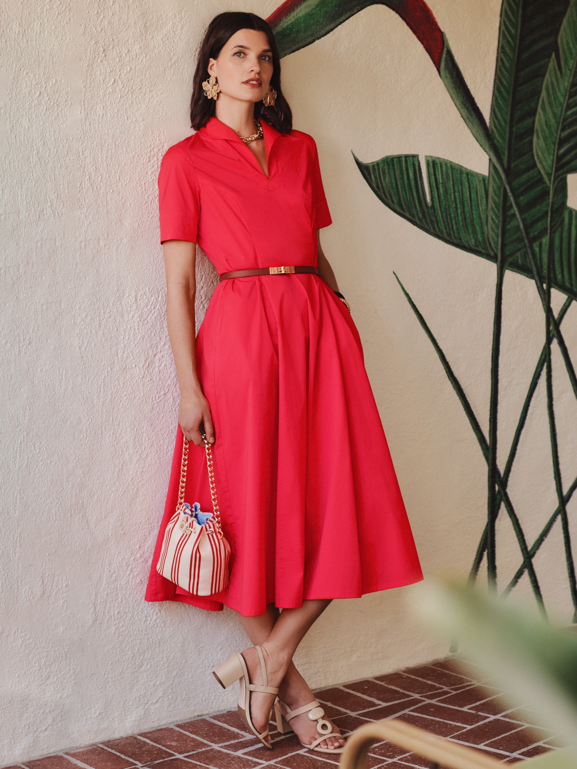 A woman wearing a vibrant red Lily Dress by J.McLaughlin, styled with a slim belt, statement earrings, a striped handbag, and nude sandals, embodying effortlessly stylish, enduring designs with a legacy of timeless fashion. A destination for defining style. Life, Well-Lived.