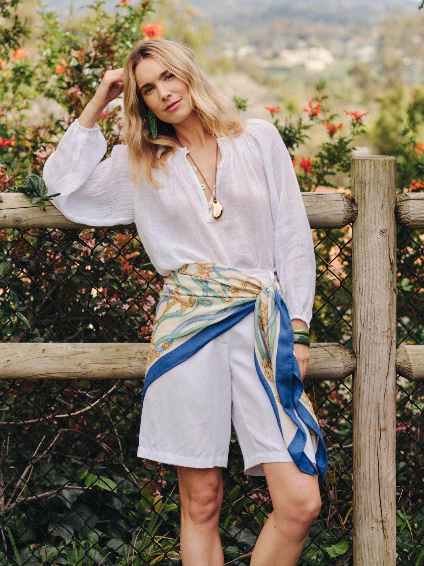 Woman stands outdoors wearing a breezy white blouse and shorts styled with the Sarita Scarf in Riding Lessons Corner by J.McLaughlin, showcasing effortlessly stylish, enduring designs and innovative fabrics.
