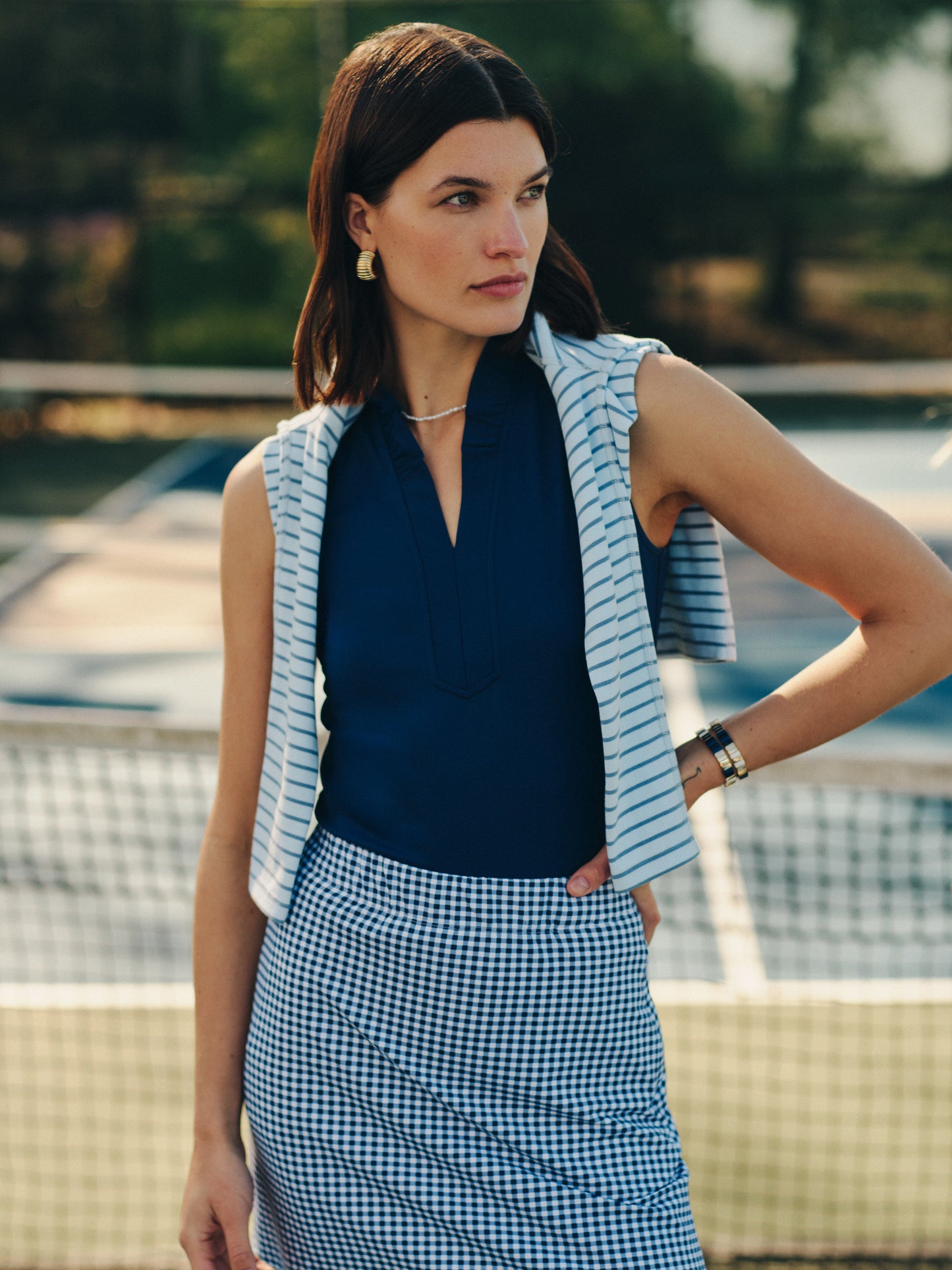 Woman wearing a navy Kate Sleeveless Ruffle Top by J.McLaughlin paired with a gingham skirt and striped sweater draped over shoulders, showcasing effortlessly stylish and enduring designs.