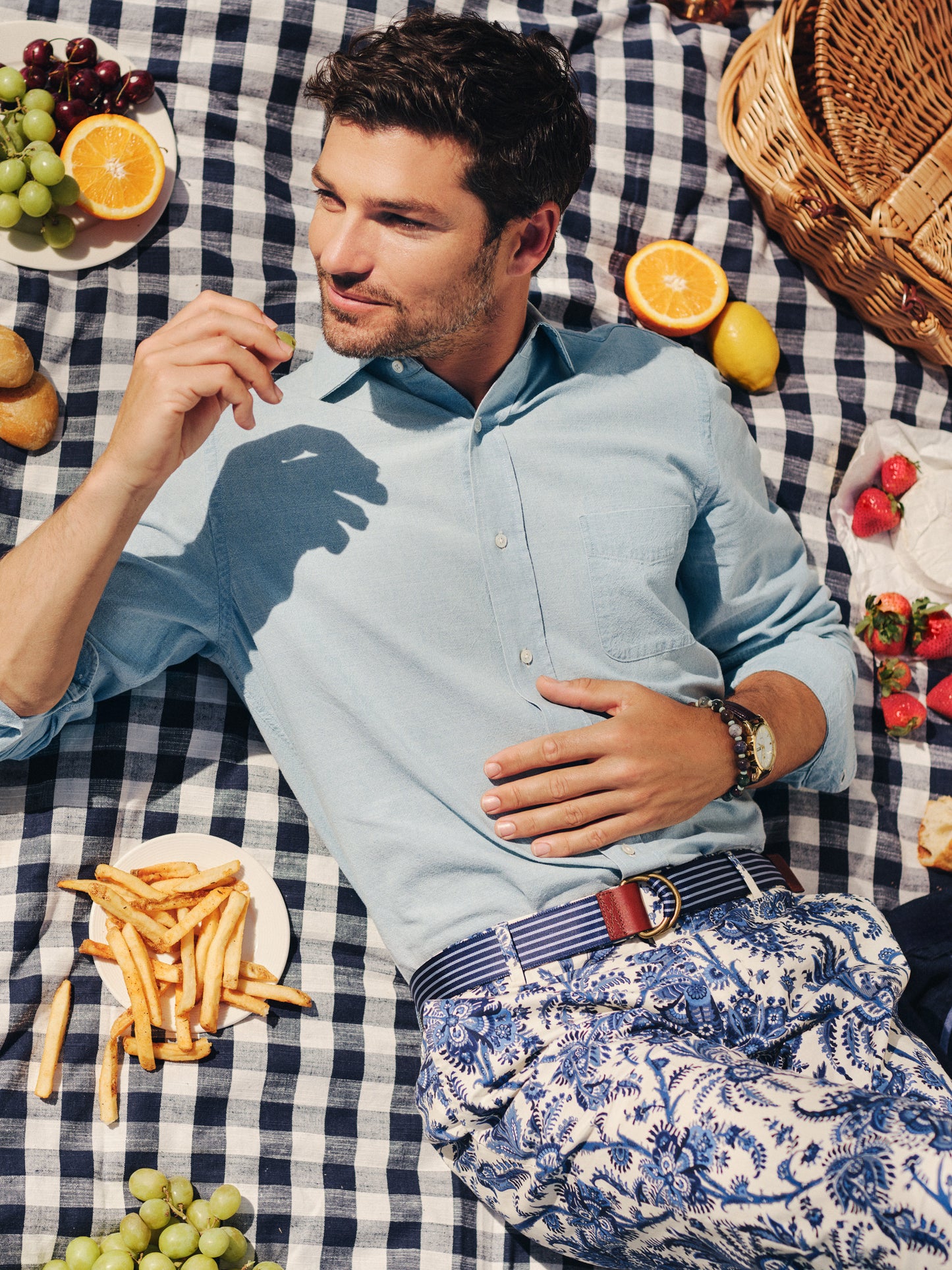 Man wearing a light blue Gramercy Cotton Chambray Shirt by J.McLaughlin lying on a checkered picnic blanket surrounded by fruits and fries, showcasing effortlessly stylish, enduring designs and innovative fabrics.