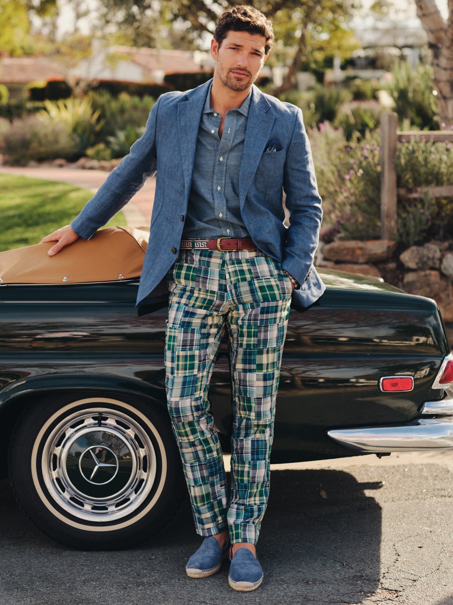 Man dressed in J.McLaughlin blue blazer and shirt paired with Madison Fancy Pants in Patchwork standing beside a classic car, showcasing effortlessly stylish, enduring designs with innovative fabrics.