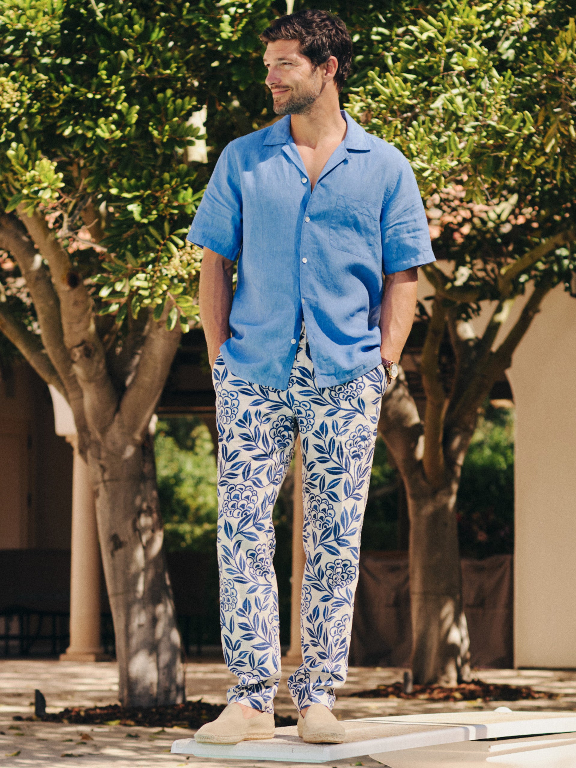 Man wearing a blue short-sleeve shirt paired with Bleeker Linen Fancy Pants in Modern Dahlia featuring a white and navy floral pattern by J.McLaughlin, showcasing effortlessly stylish, enduring designs and innovative fabrics. A destination for defining style. Life, Well-Lived.