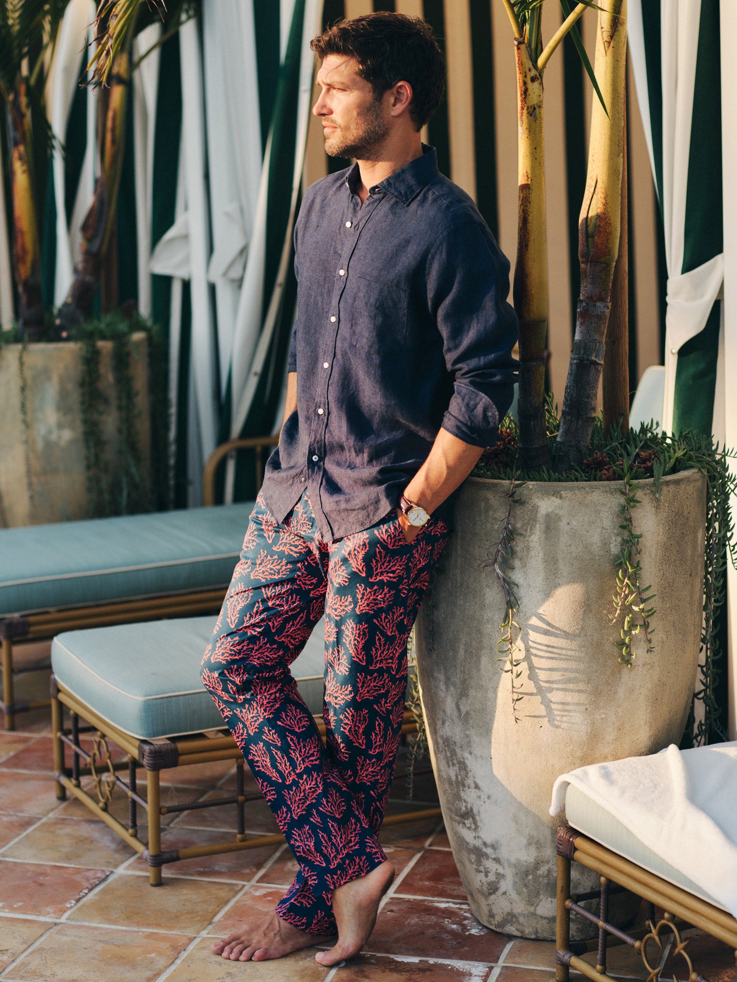 Man wearing a navy shirt and Madison Fancy Pants in Midi Coral Cay Texture with a coral pattern, standing barefoot next to a large planter outdoors, showcasing J.McLaughlin’s effortlessly stylish, enduring designs and innovative fabrics. A destination for defining style. Life, Well-Lived.