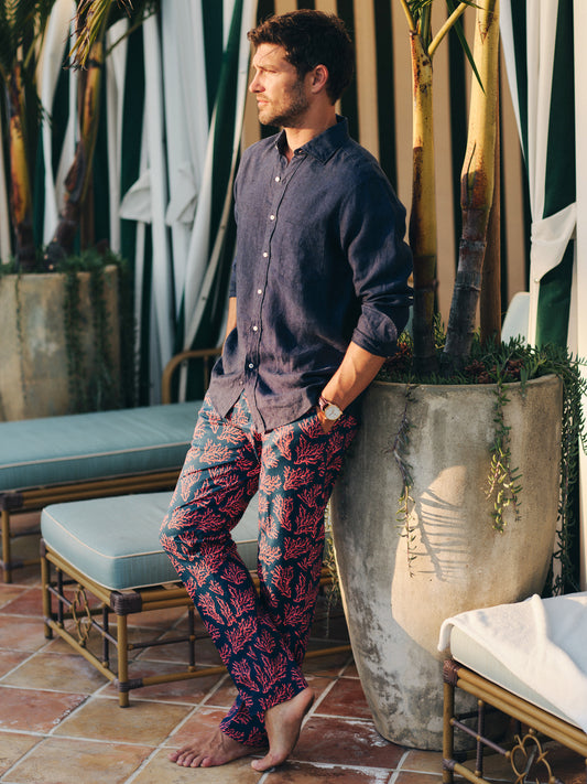 Man wearing a navy shirt and Madison Fancy Pants in Midi Coral Cay Texture with a coral pattern, standing barefoot next to a large planter outdoors, showcasing J.McLaughlin’s effortlessly stylish, enduring designs and innovative fabrics. A destination for defining style. Life, Well-Lived.