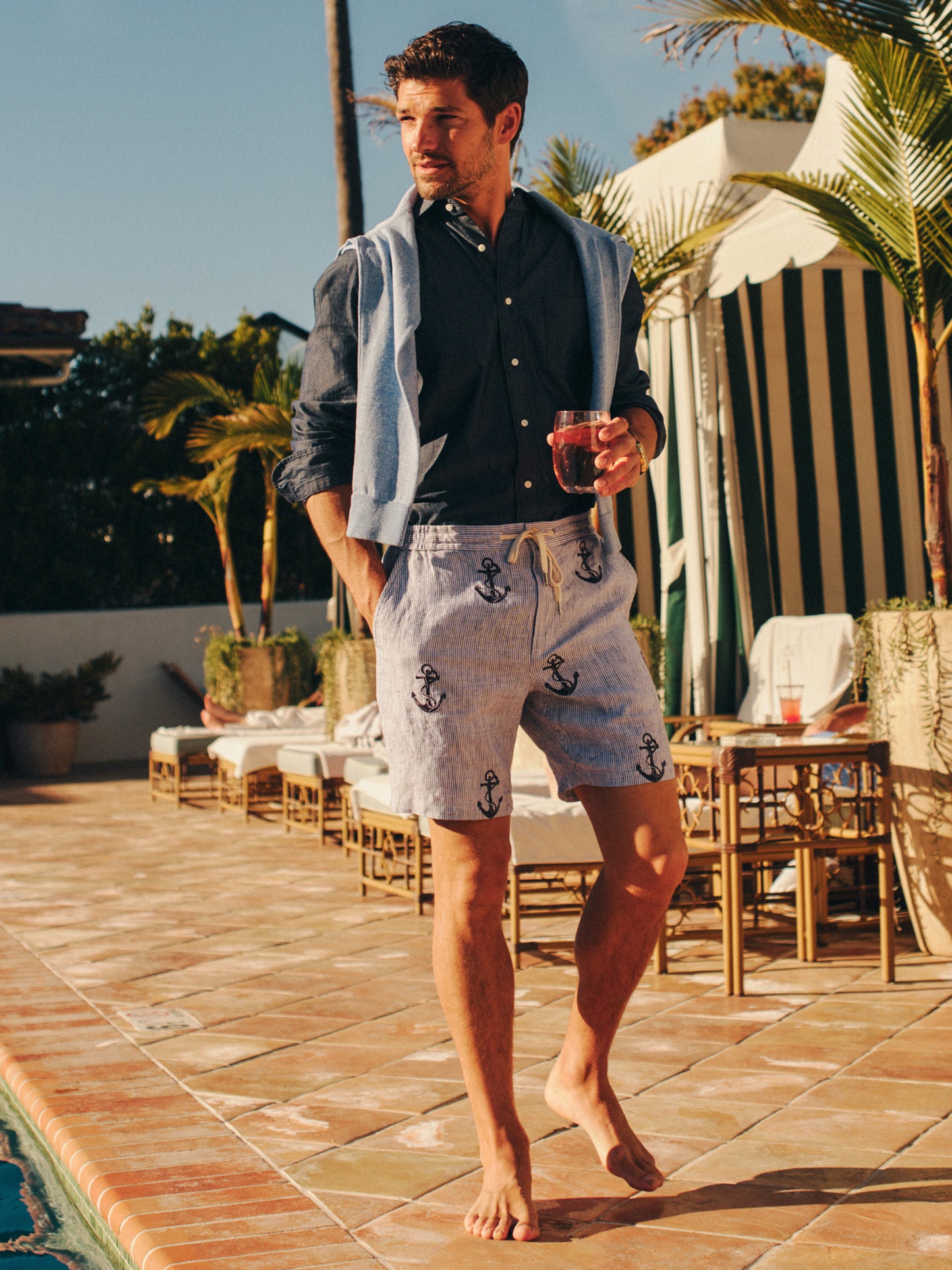 Man by poolside wearing Newport Linen Shorts in Anchor by J.McLaughlin with a navy shirt and light sweater draped over shoulders, embodying effortlessly stylish, enduring designs in innovative fabrics.
