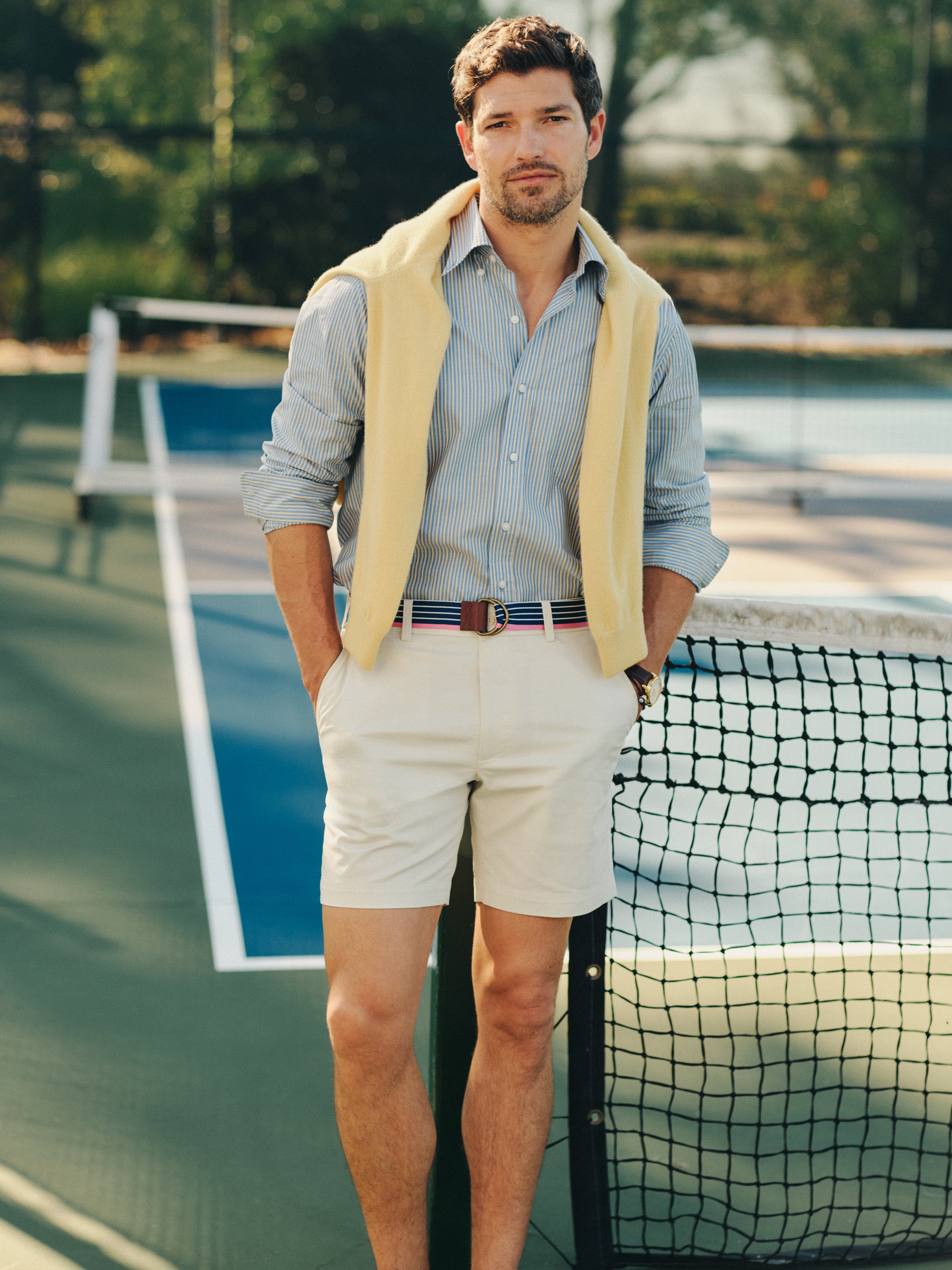Man wearing J.McLaughlin Chatham Shorts paired with a striped shirt and yellow sweater draped over shoulders, embodying effortlessly stylish, enduring designs in a classic tennis court setting.
