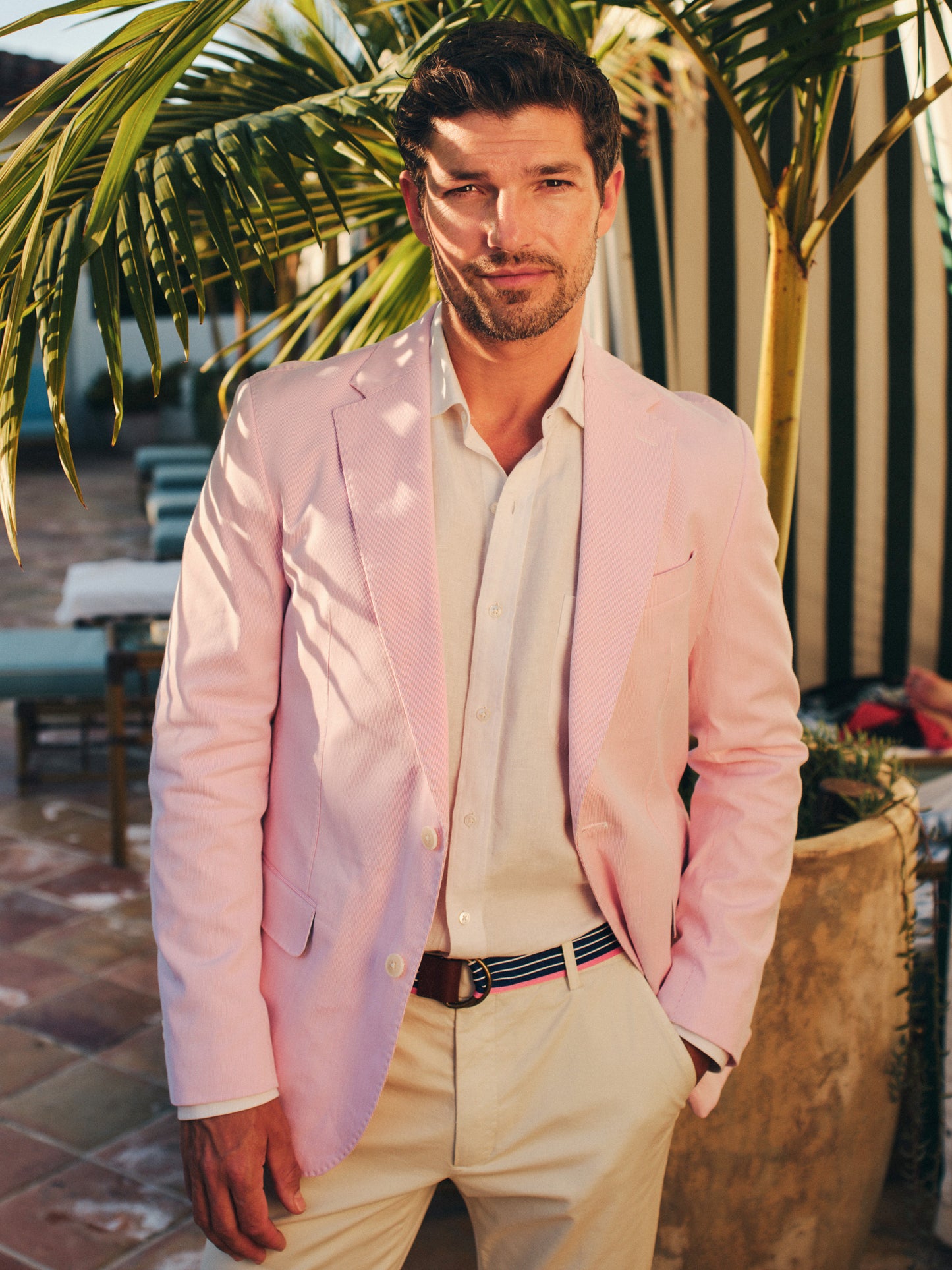 Man wearing a Suffolk Sport Coat in Fineline by J.McLaughlin in soft pink paired with a light shirt and light trousers, standing outdoors near tropical plants, effortlessly stylish, enduring designs, innovative fabrics, legacy of timeless fashion. A destination for defining style. Life, Well-Lived.