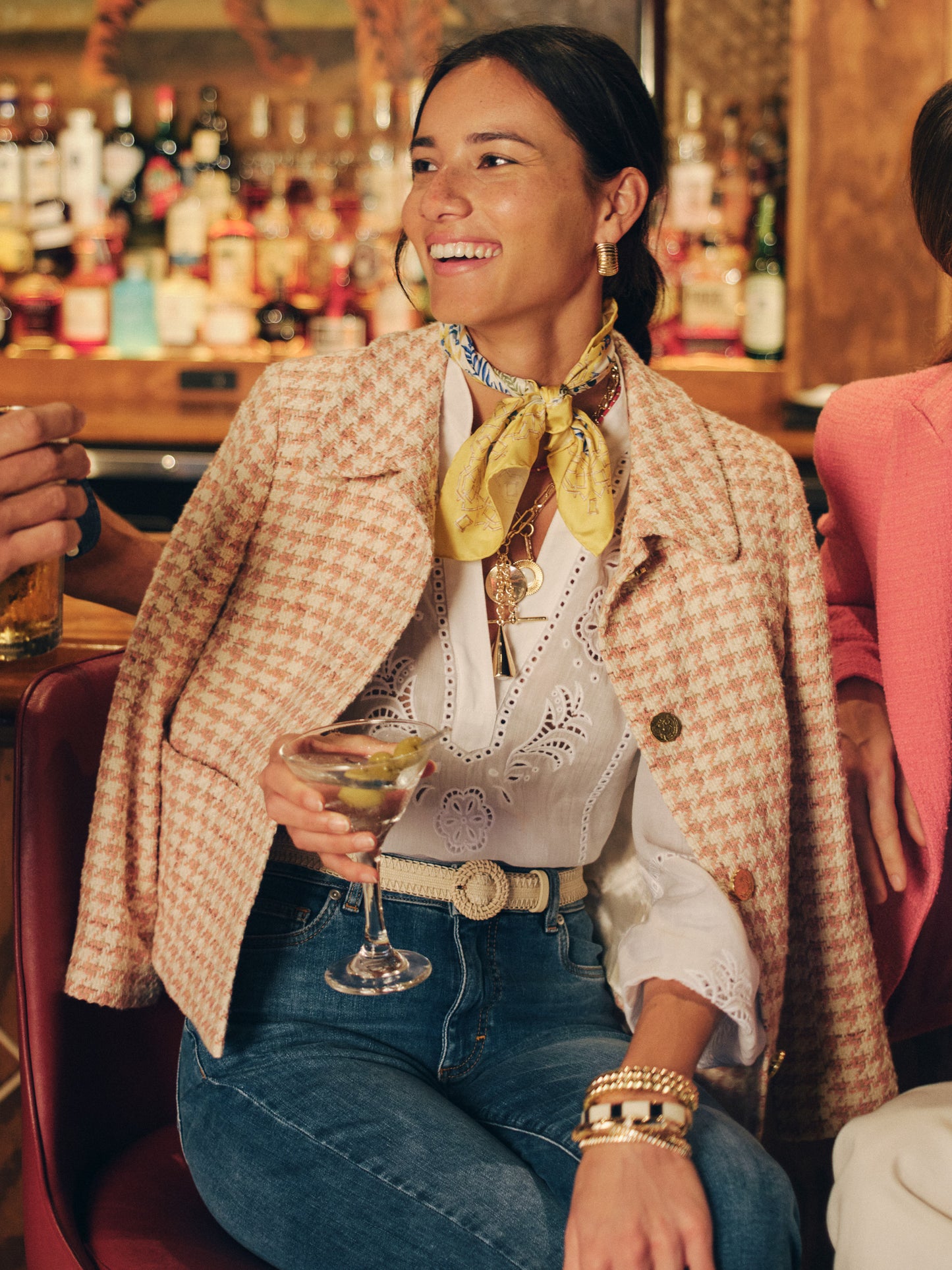 Woman wearing a Violette Tweed Jacket in Houndstooth by J.McLaughlin over a white eyelet blouse, accessorized with a yellow scarf and bracelets, smiling while holding a cocktail, showcasing effortlessly stylish, enduring designs and innovative fabrics in a lively setting.