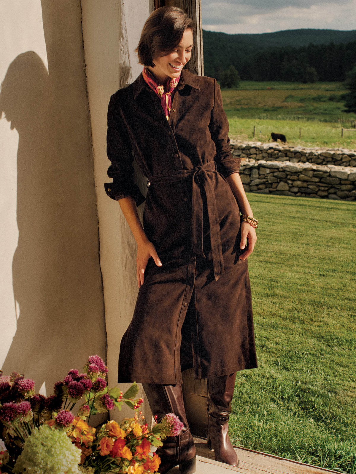 J.McLaughlin Amory Suede Maxi Dress in rich chocolate brown with shirt dress styling and a tie waist worn outdoors beside a stone wall