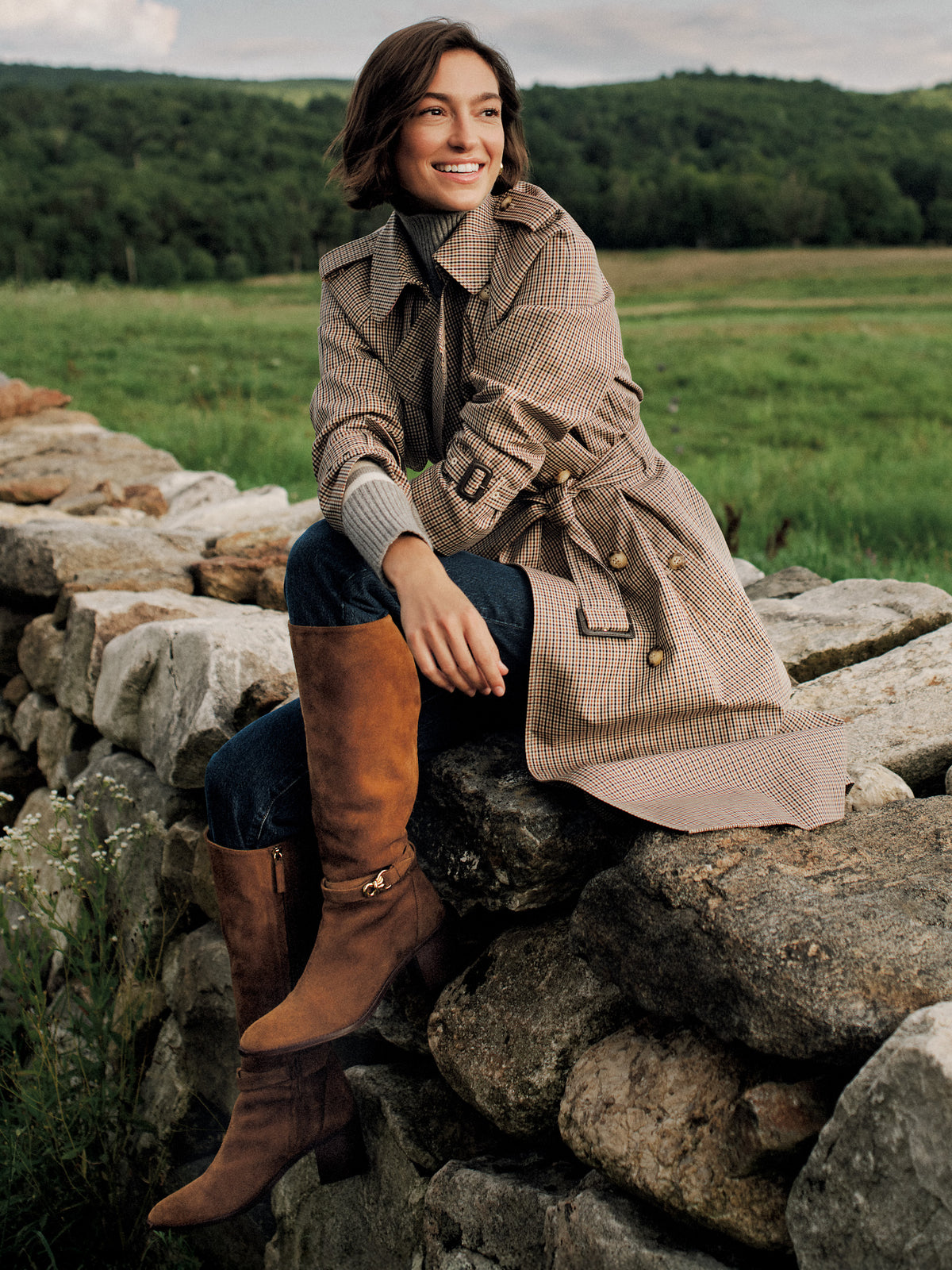 Campbell Trench Coat in Check by J.McLaughlin worn with tall brown boots by a woman seated on rocks in a scenic countryside setting
