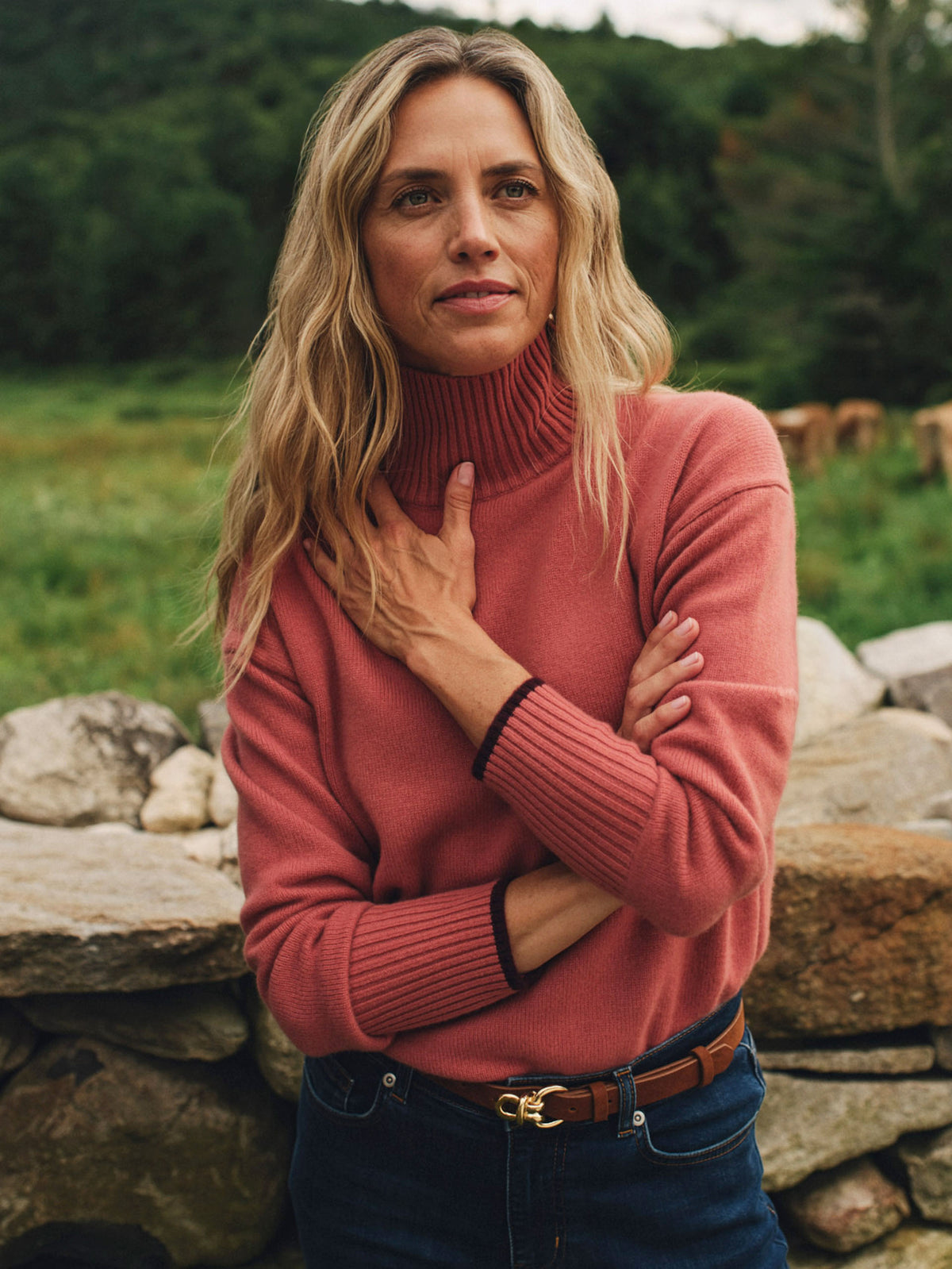Woman wearing the Dina Cashmere Turtleneck by J.McLaughlin in coral with dark jeans and a leather belt outdoors among stone walls and greenery