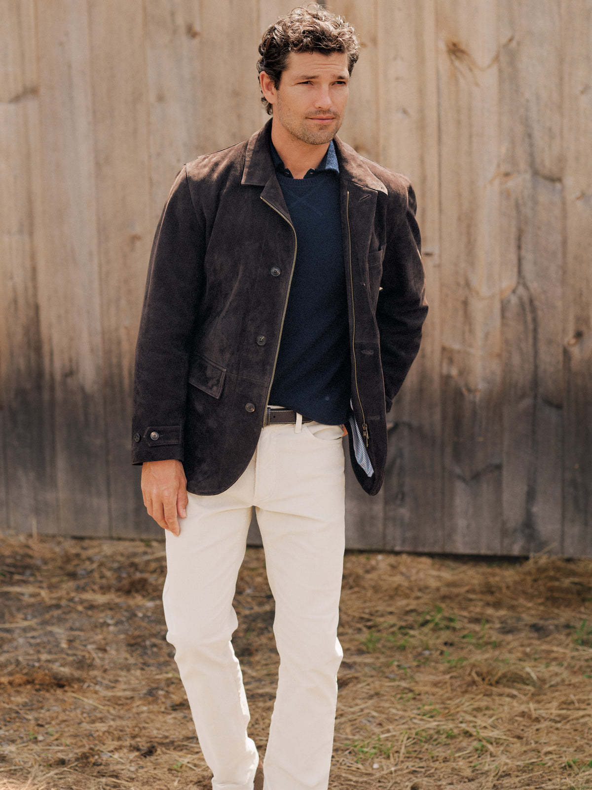 J.McLaughlin Winston Suede Jacket in dark brown suede worn with a navy sweater and cream pants by a man outdoors beside a rustic wooden wall