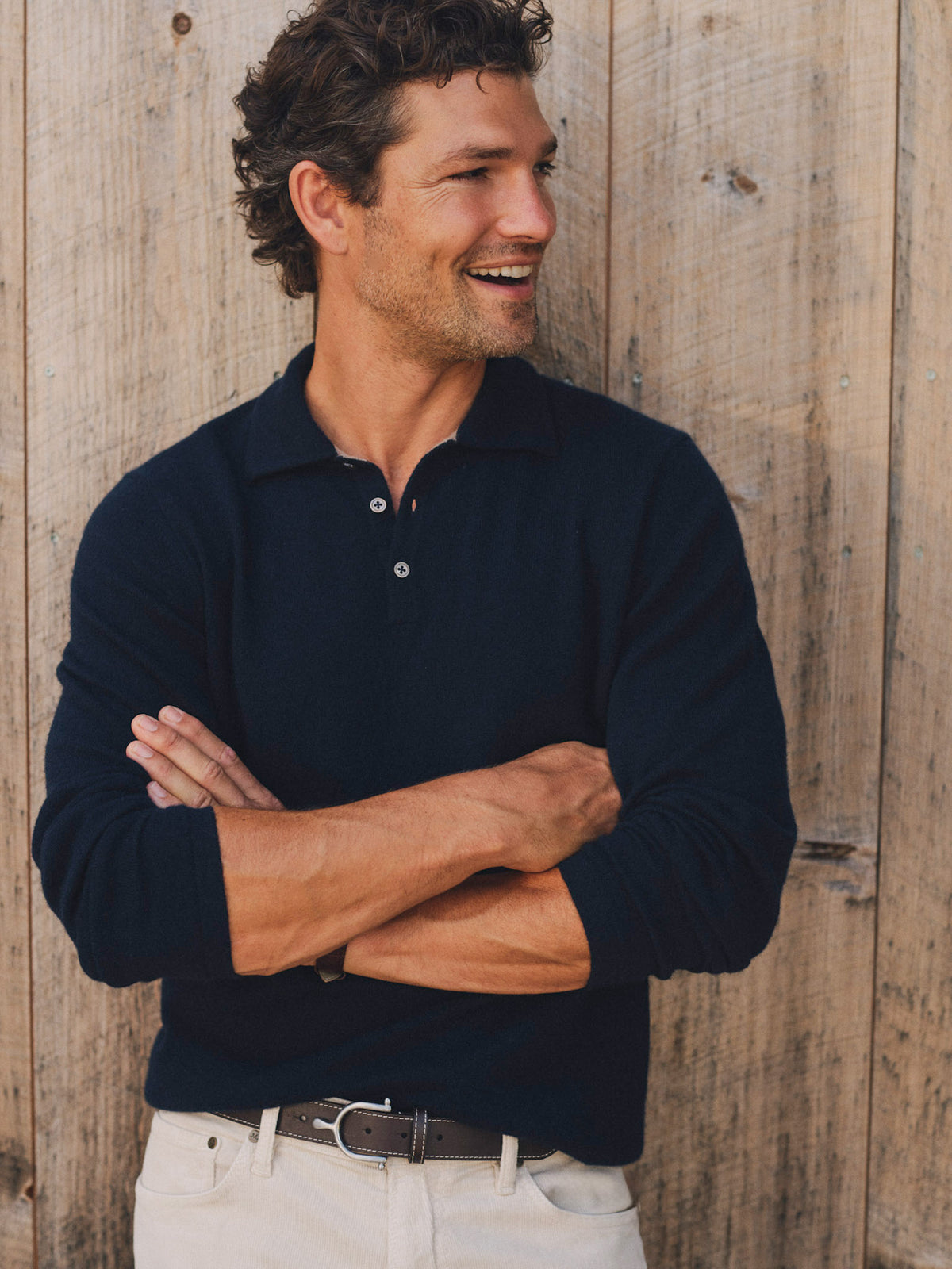 Smiling man wearing the Langston Cashmere Polo Sweater by J.McLaughlin with crossed arms against a rustic wood wall