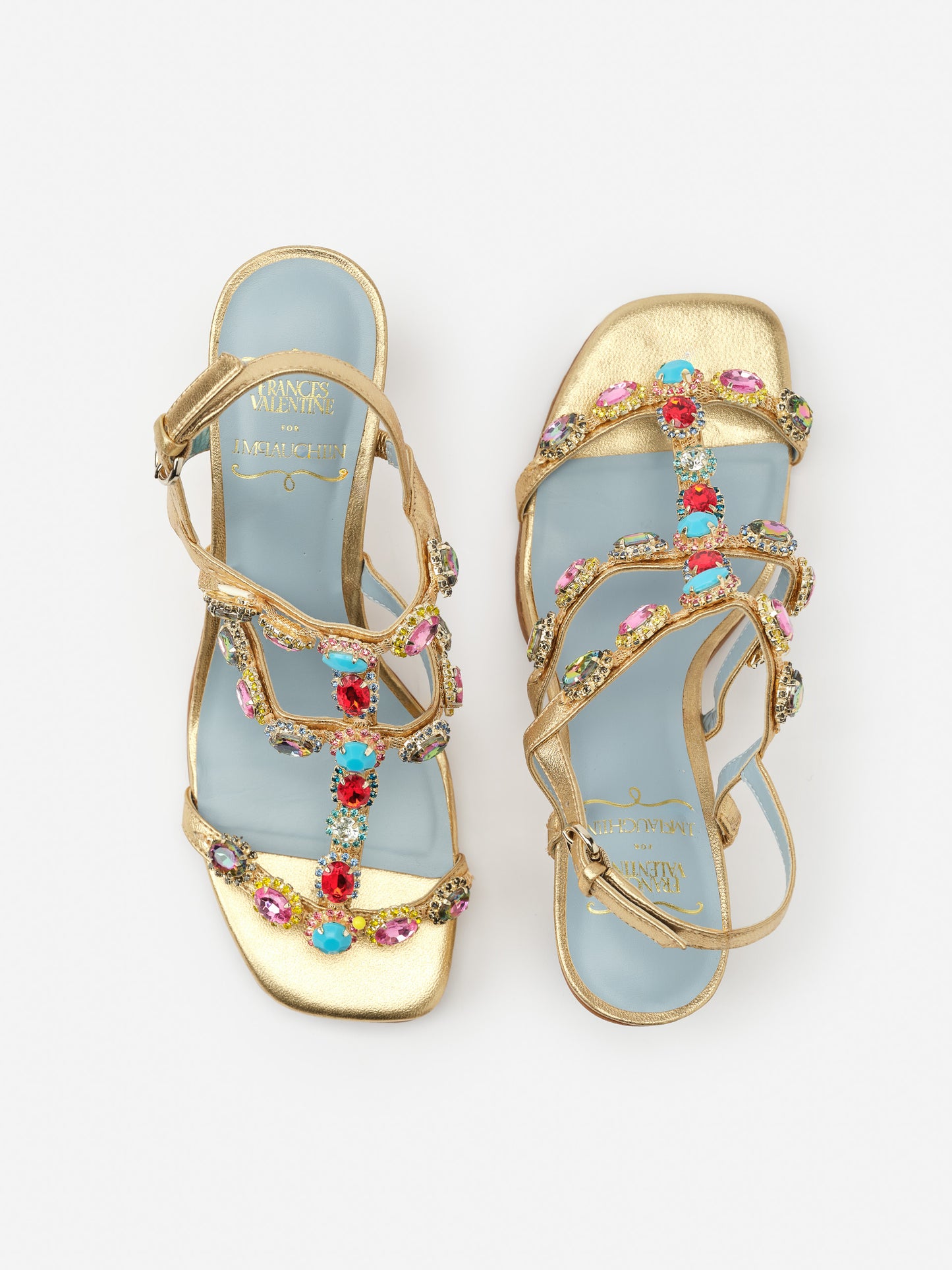 Kiki Gem Heels by J.McLaughlin feature gold straps adorned with colorful gemstone embellishments on a light blue sole, blending effortlessly stylish design with enduring appeal.