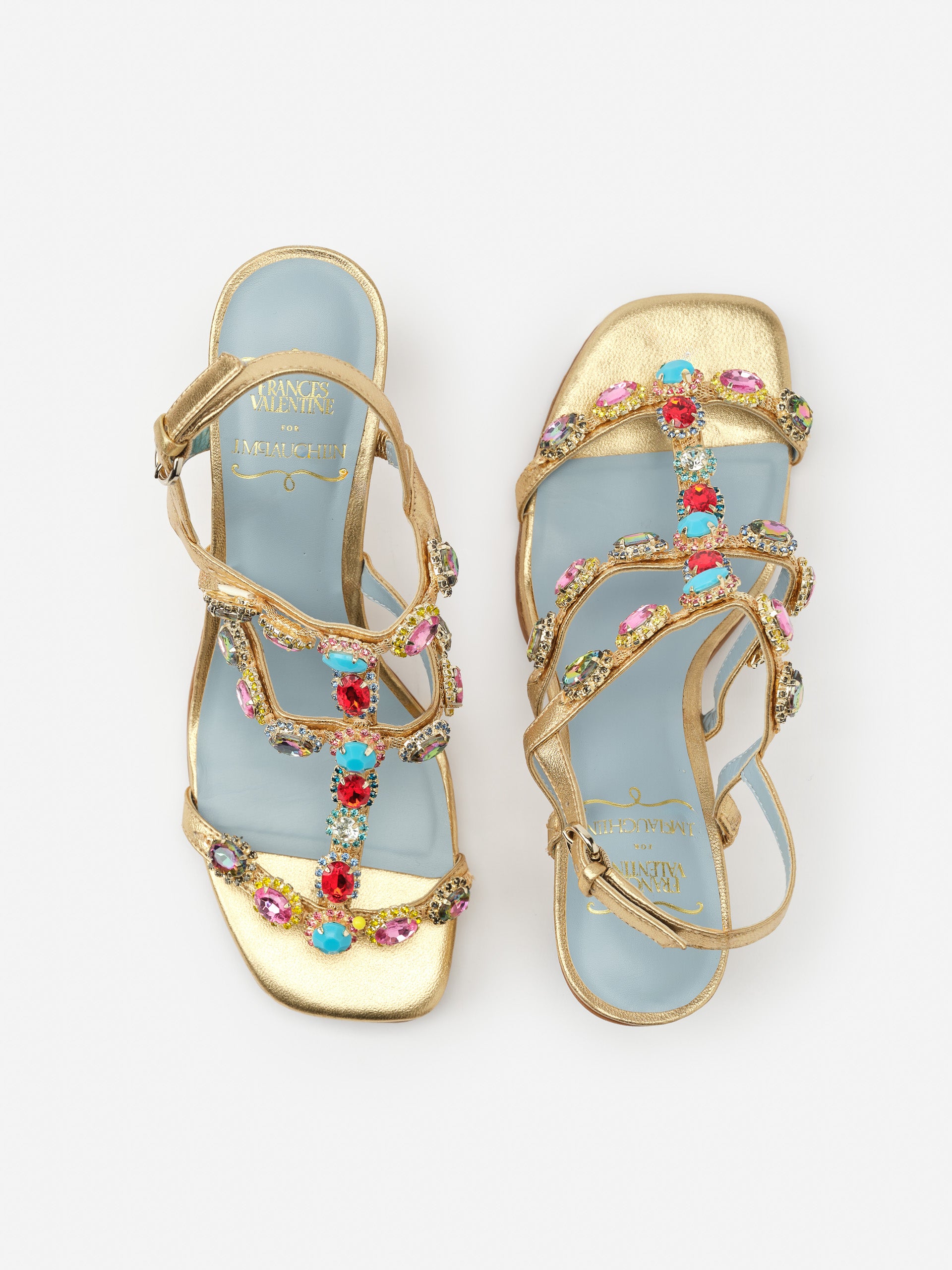 Kiki Gem Heels by J.McLaughlin feature gold straps adorned with colorful gemstone embellishments on a light blue sole, blending effortlessly stylish design with enduring appeal.