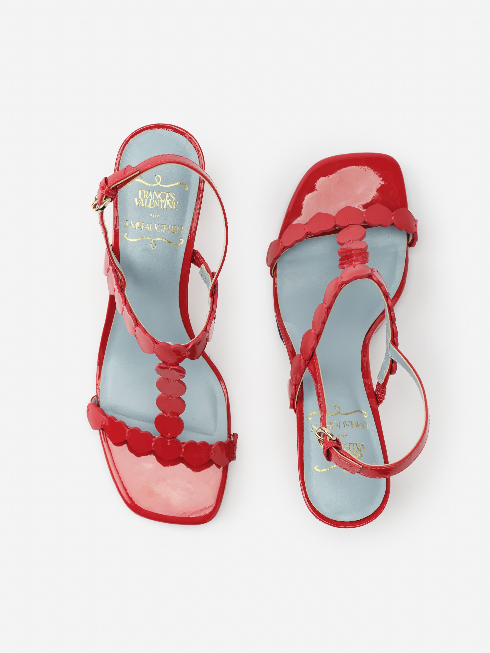 Pair of glossy red Kiki Heart Heels by J.McLaughlin with heart-shaped embellishments and ankle straps, embodying effortlessly stylish, enduring designs and a legacy of timeless fashion.