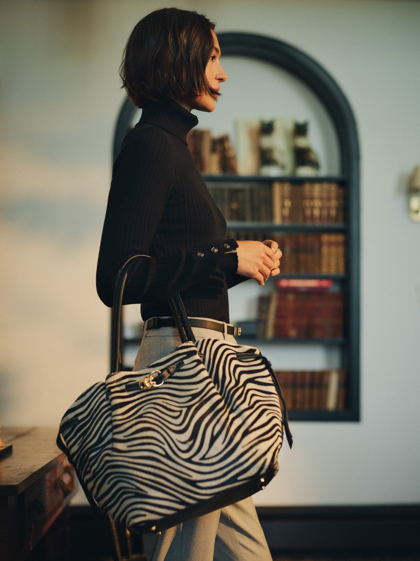 Woman wearing a black turtleneck and beige pants holding The Lex Pony Hair Tote Bag in Zebra Stripes by J.McLaughlin, showcasing an effortlessly stylish and timeless patterned bag.