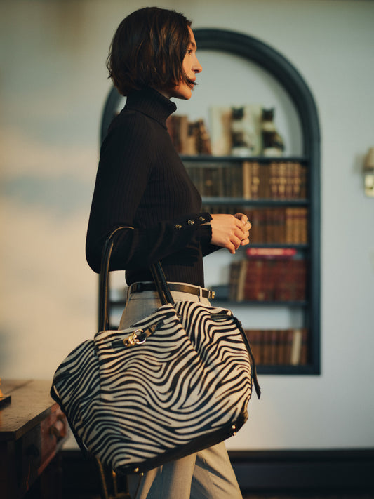 Woman wearing a black turtleneck and beige pants holding The Lex Pony Hair Tote Bag in Zebra Stripes by J.McLaughlin, showcasing an effortlessly stylish and timeless patterned bag.