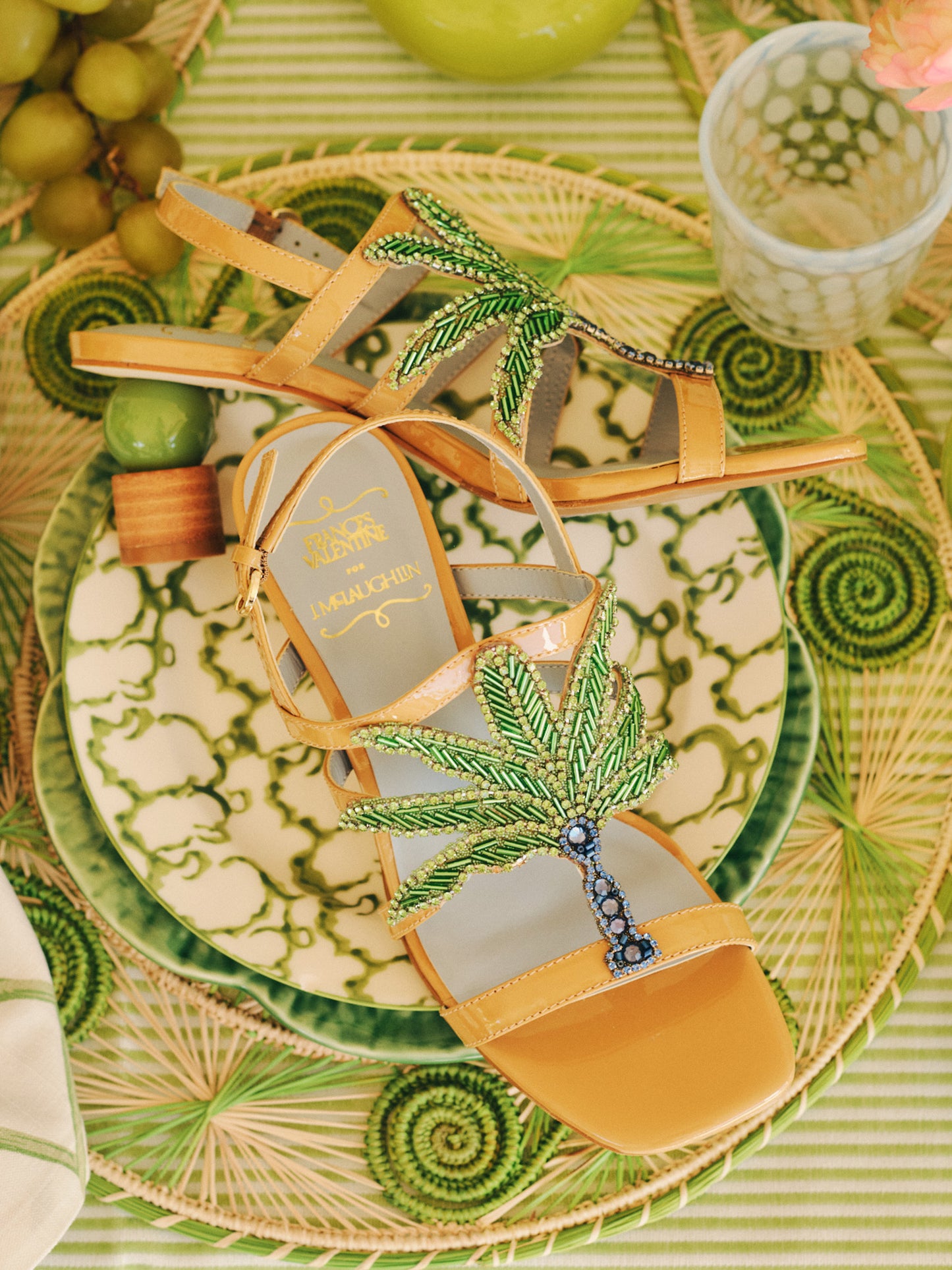Venus Palm Tree Heels by J.McLaughlin feature intricate green palm tree beadwork on tan leather sandals, displayed on patterned green and beige plates with a stylish summer vibe.
