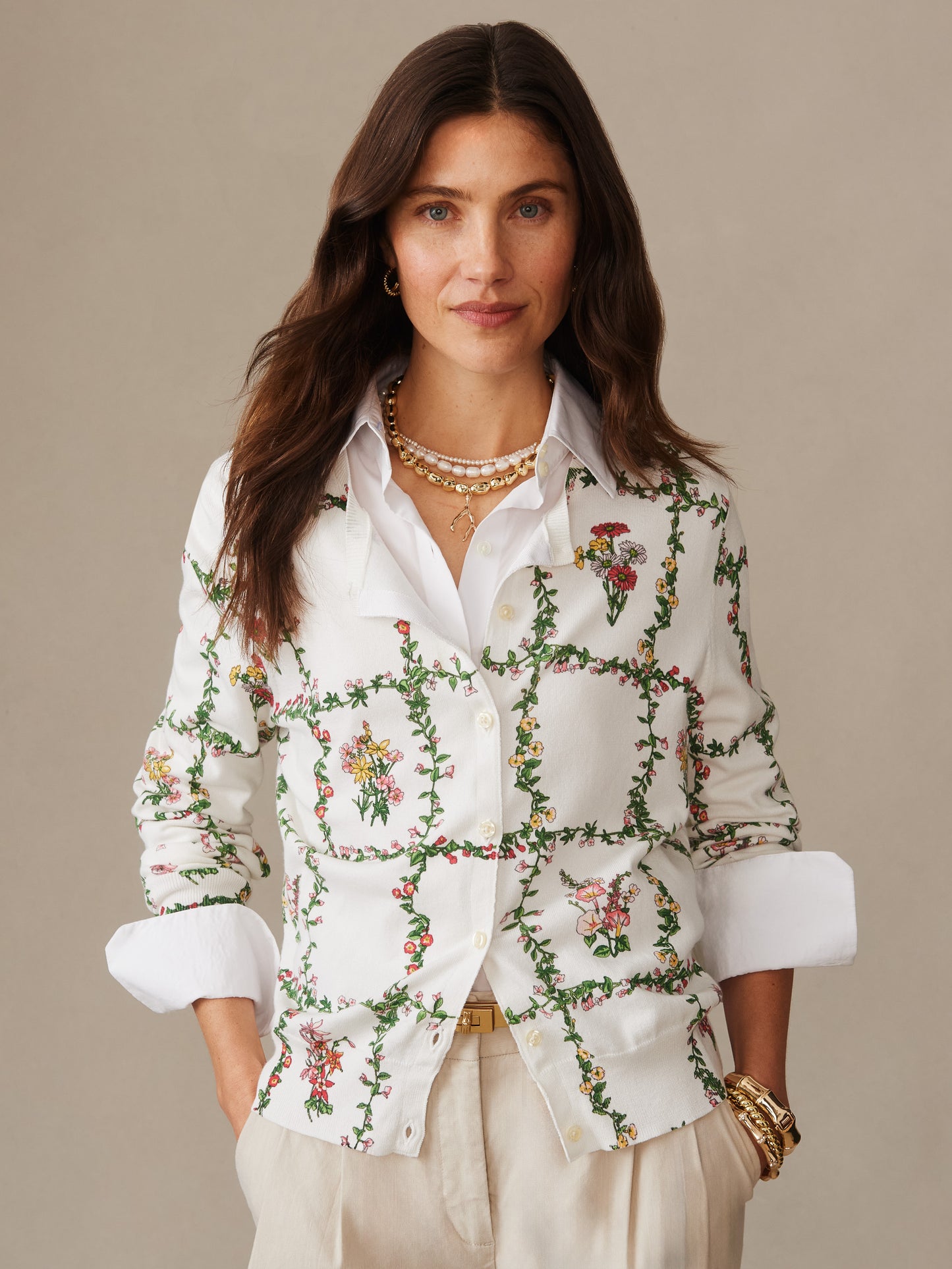 Woman wearing a Whitney Cardigan in Floral Medley Frame by J.McLaughlin, featuring a white floral patterned sweater layered over a white shirt, styled with beige pants and gold accessories, showcasing effortlessly stylish, enduring designs and a legacy of timeless fashion.