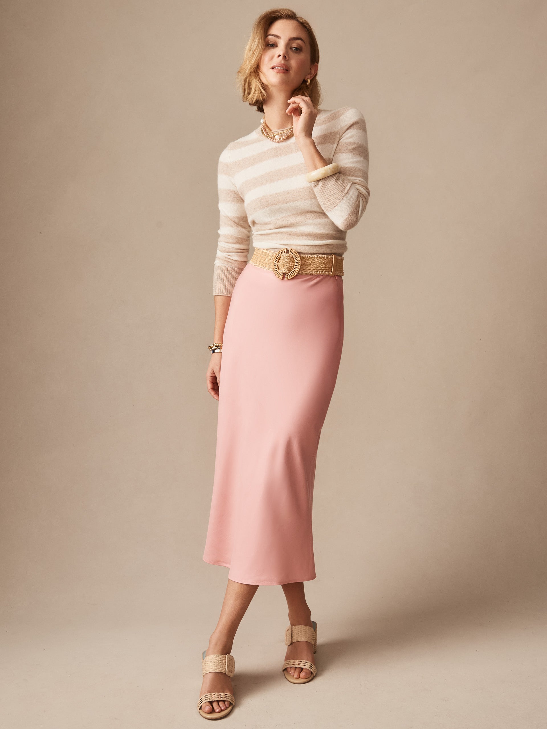 A woman wears the Adelyn Skirt in soft pink paired with a beige striped top, tan belt, and sandals, showcasing J.McLaughlin’s legacy of timeless fashion with effortlessly stylish, enduring designs.