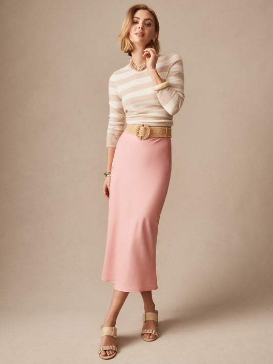 A woman wears the Adelyn Skirt in soft pink paired with a beige striped top, tan belt, and sandals, showcasing J.McLaughlin’s legacy of timeless fashion with effortlessly stylish, enduring designs.