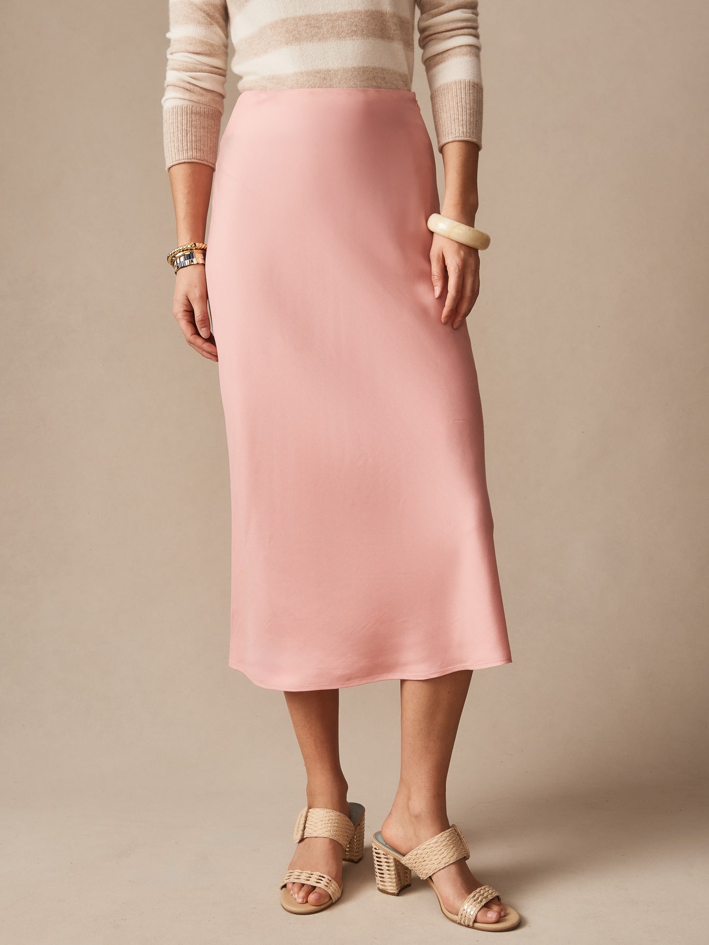 Model wearing a softly draped pink Adelyn Skirt by J.McLaughlin, paired with neutral striped top and woven heeled sandals, showcasing effortlessly stylish, enduring designs.