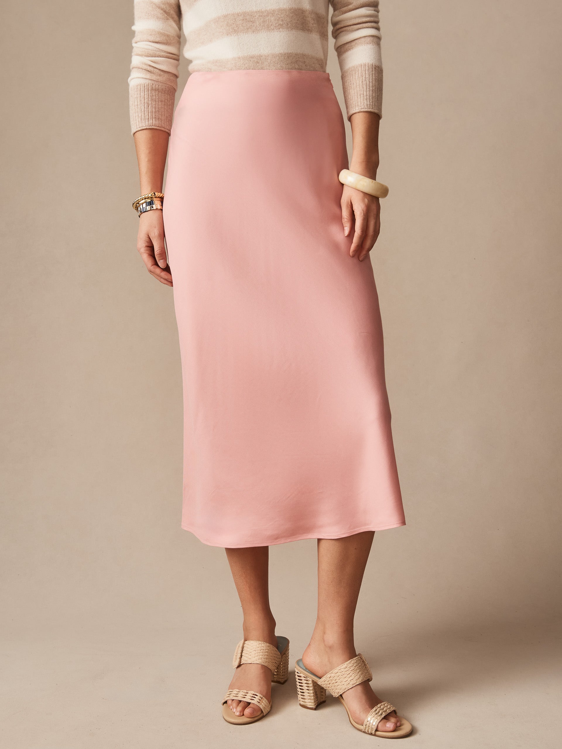 Model wearing a softly draped pink Adelyn Skirt by J.McLaughlin, paired with neutral striped top and woven heeled sandals, showcasing effortlessly stylish, enduring designs.
