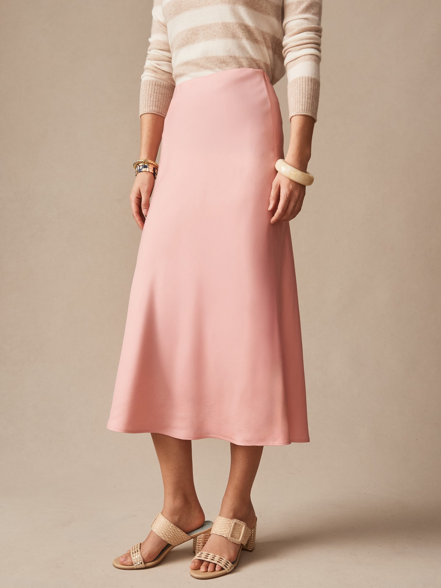 Woman wearing a soft pink Adelyn Skirt by J.McLaughlin paired with a striped beige sweater and woven sandals, showcasing effortlessly stylish, enduring designs in innovative fabrics.