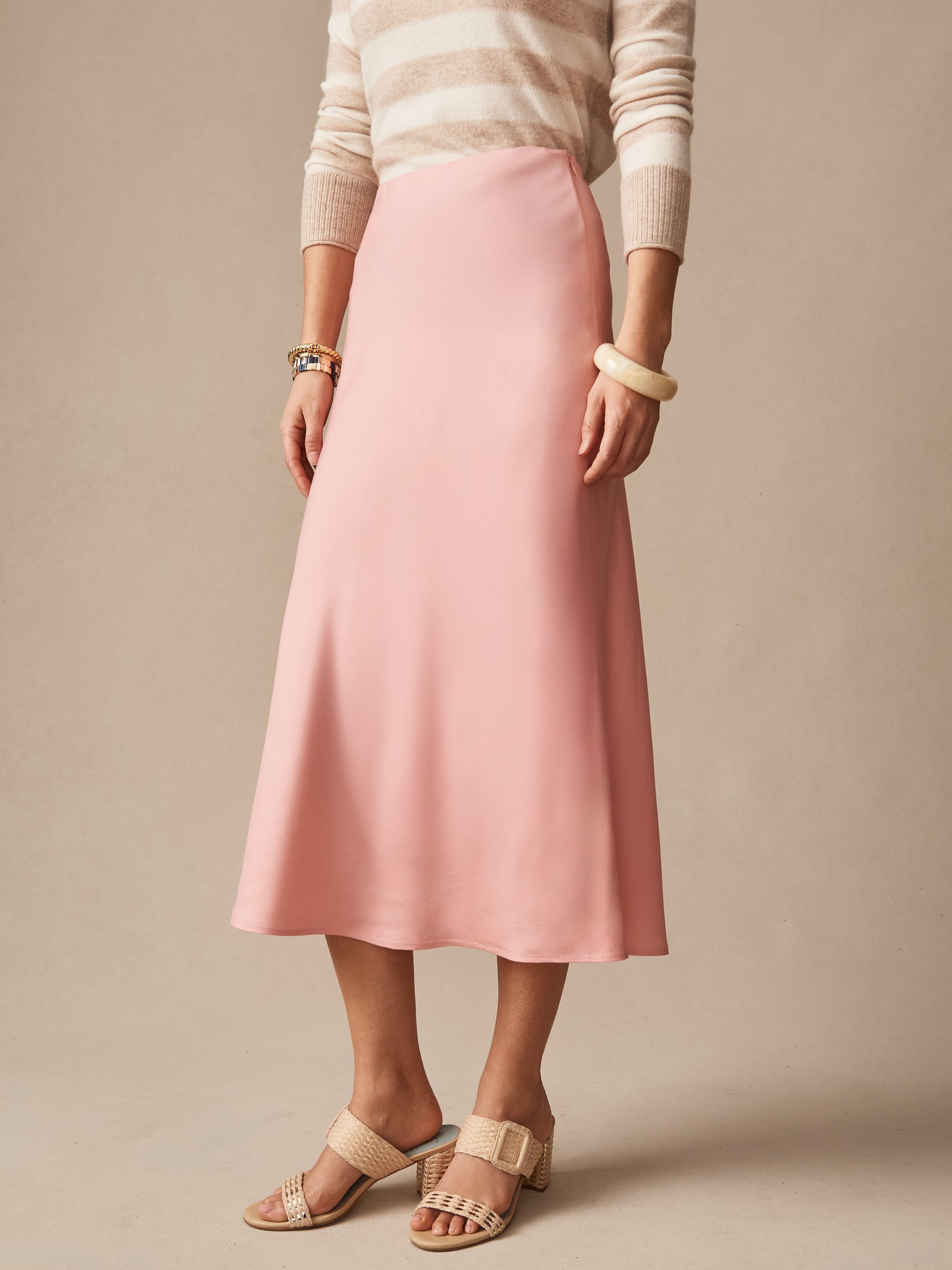 Woman wearing a soft pink Adelyn Skirt by J.McLaughlin paired with a striped beige sweater and woven sandals, showcasing effortlessly stylish, enduring designs in innovative fabrics.