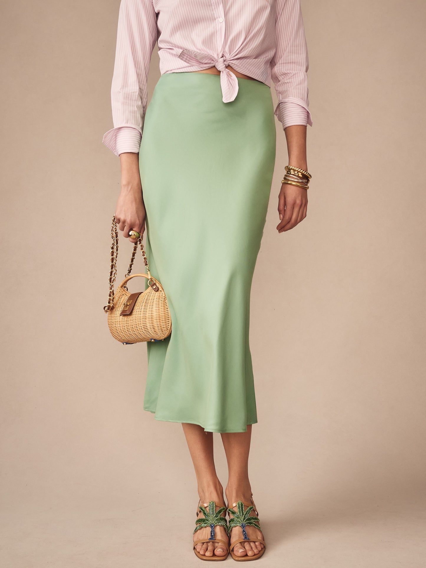 Woman wearing a soft green Adelyn Skirt by J.McLaughlin paired with a pink tied blouse, accessorized with a woven handbag and stylish sandals, showcasing effortlessly stylish, enduring designs and innovative fabrics in a legacy of timeless fashion. A destination for defining style. Life, Well-Lived.