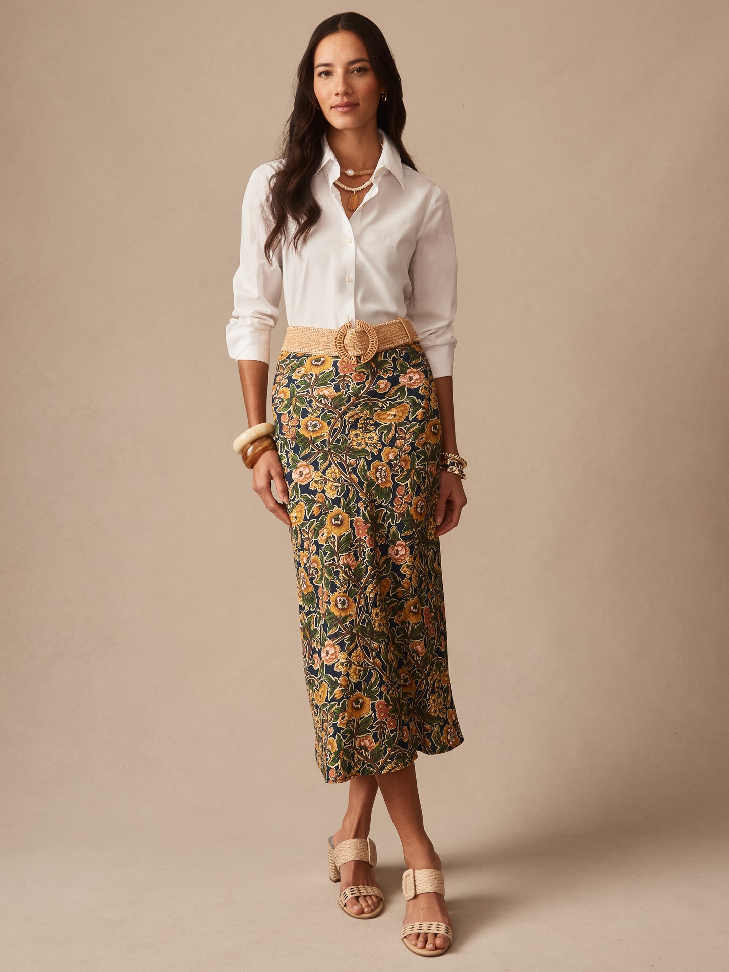 A woman modeling the Adelyn Skirt in Bucolic Blooms Bias by J.McLaughlin, paired with a white shirt, neutral woven belt, and sandals, showcasing effortlessly stylish, enduring designs that reflect a legacy of timeless fashion. A destination for defining style. Life, Well-Lived.