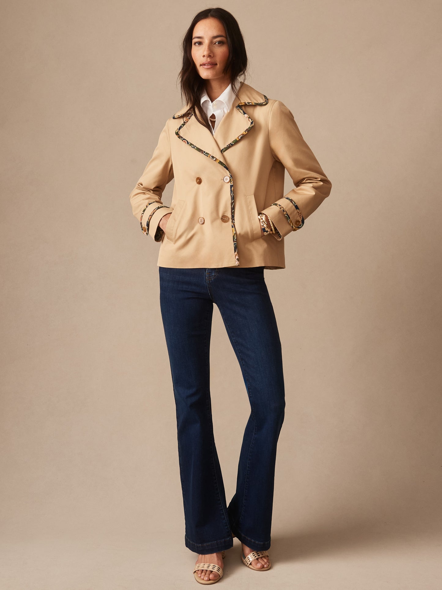 Woman wearing a beige Audrey Jacket by J.McLaughlin featuring patterned trim, paired with dark blue jeans and sandals, showcasing effortlessly stylish, enduring designs.