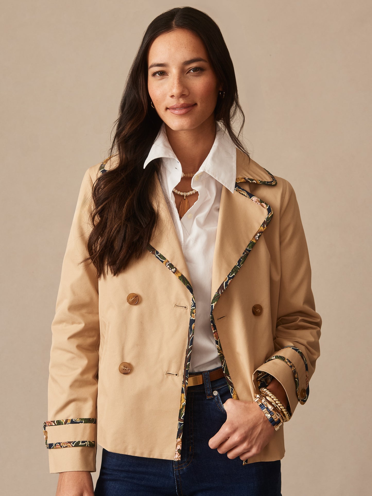 Woman wearing a beige Audrey Jacket by J.McLaughlin featuring floral trim details, paired with a white shirt and dark jeans, showcasing effortlessly stylish, enduring designs and innovative fabrics.