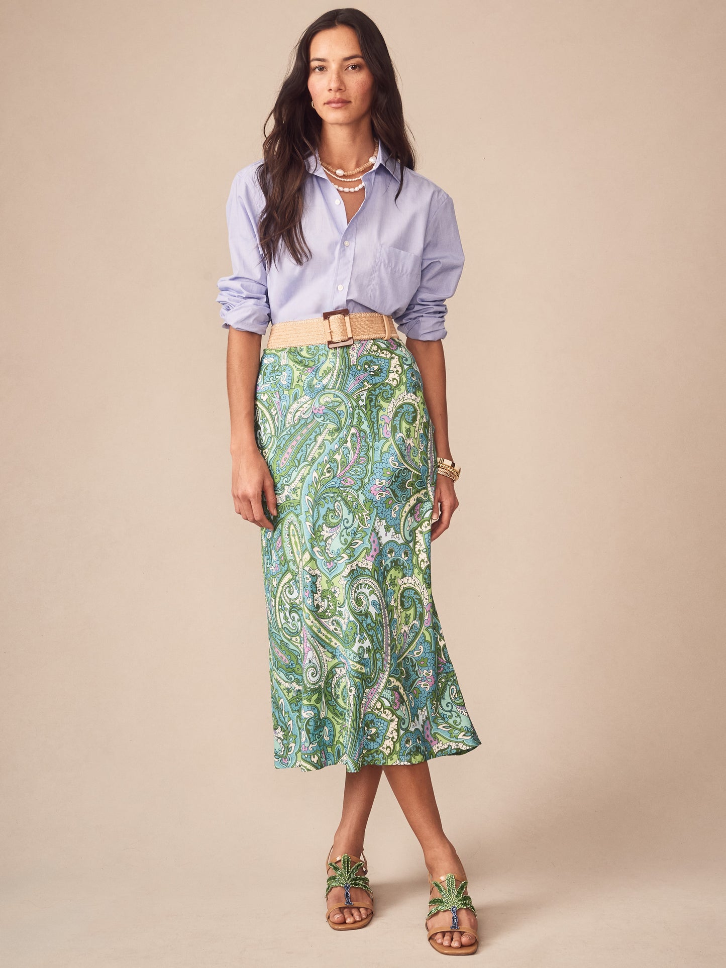 A woman wearing a light blue button-down shirt paired with the Adelyn Skirt in Paisley Reverie Bias by J.McLaughlin featuring a green paisley pattern, styled with a beige woven belt and sandals, showcasing effortlessly stylish, enduring designs with innovative fabrics, a destination for defining style, Life, Well-Lived.