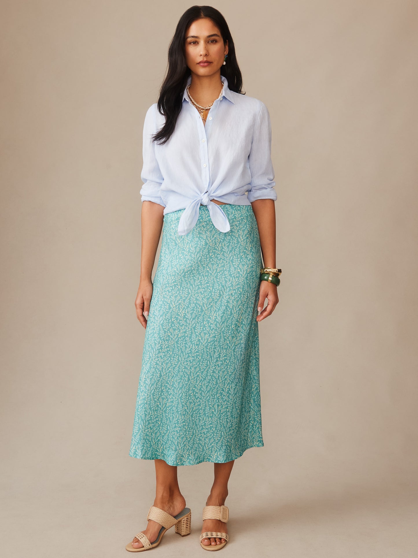Woman wearing a light blue button-up shirt tied at the waist paired with an Adelyn Silk Blend Skirt in Mini Sea Vine Bias in teal with subtle vine pattern, styled with neutral sandals and layered jewelry, showcasing J.McLaughlin’s effortlessly stylish, enduring designs and innovative fabrics in a legacy of timeless fashion. A destination for defining style. Life, Well-Lived.