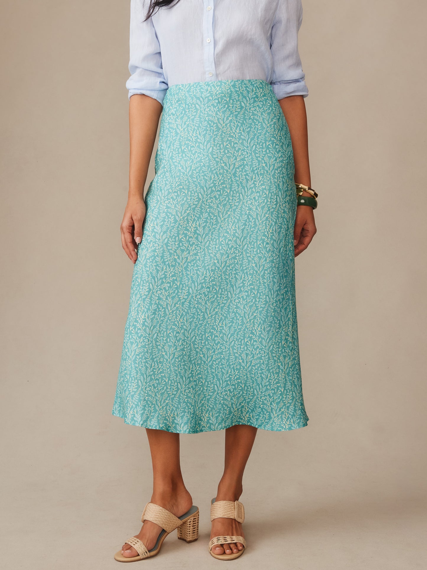 Woman wearing a light blue button-up shirt paired with the Adelyn Silk Blend Skirt in Mini Sea Vine Bias by J.McLaughlin, featuring a delicate sea vine pattern on a teal background, styled with beige open-toe heels and bracelets. Effortlessly stylish and enduring designs highlight innovative fabrics in a legacy of timeless fashion. A destination for defining style. Life, Well-Lived.