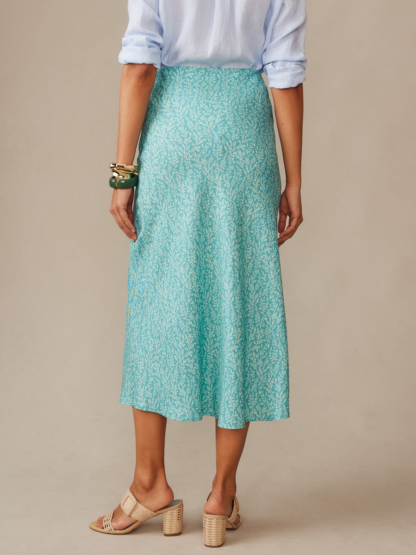 Woman wearing a teal patterned Adelyn Silk Blend Skirt in Mini Sea Vine Bias by J.McLaughlin, paired with a light blue blouse and beige heeled sandals, showcasing effortlessly stylish enduring designs with innovative fabrics and a legacy of timeless fashion. A destination for defining style. Life, Well-Lived.