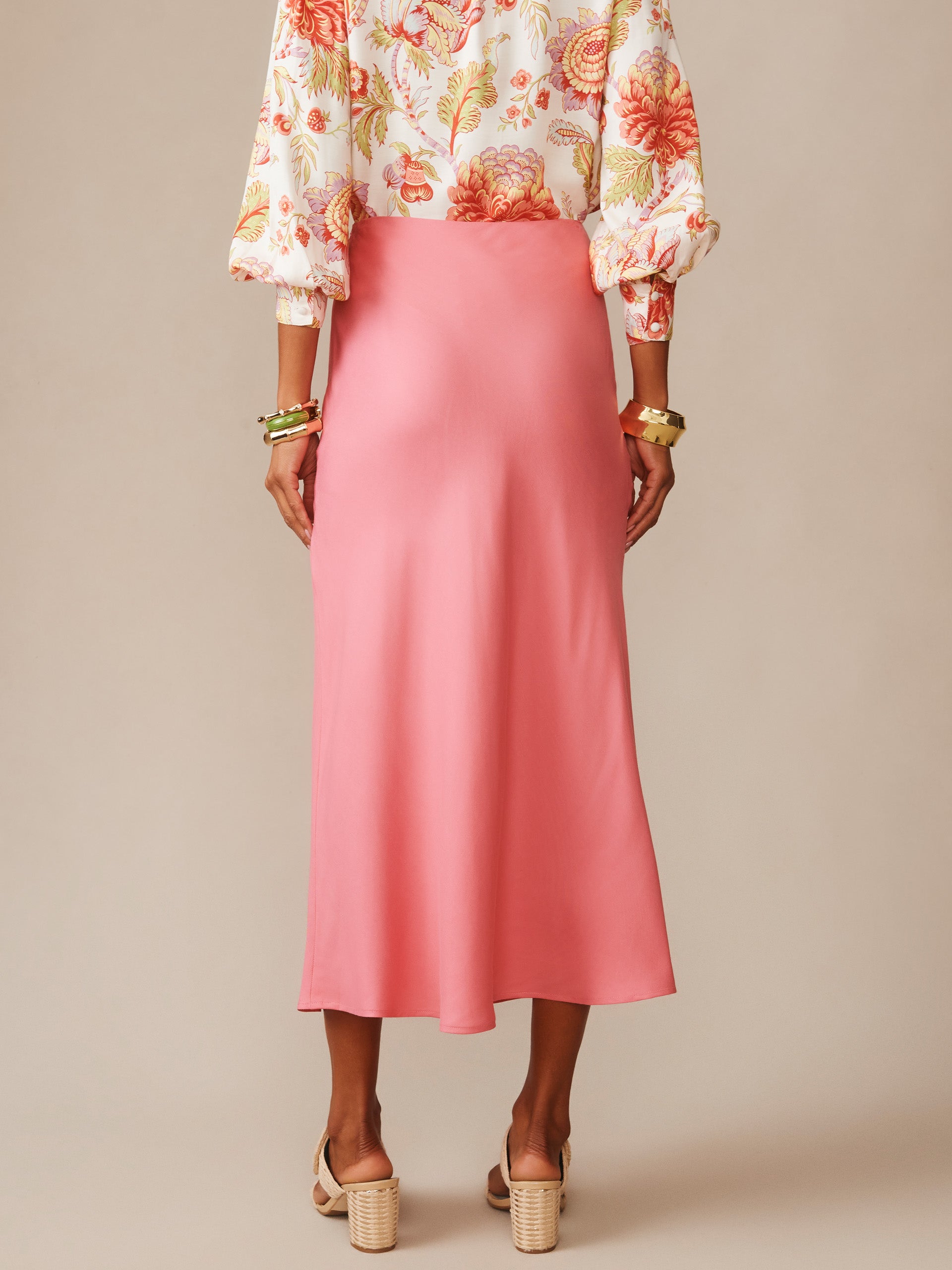 Woman wearing a soft coral Adelyn Silk Blend Skirt by J. McLaughlin, paired with a floral blouse and heeled sandals, showcasing effortlessly stylish, enduring designs.