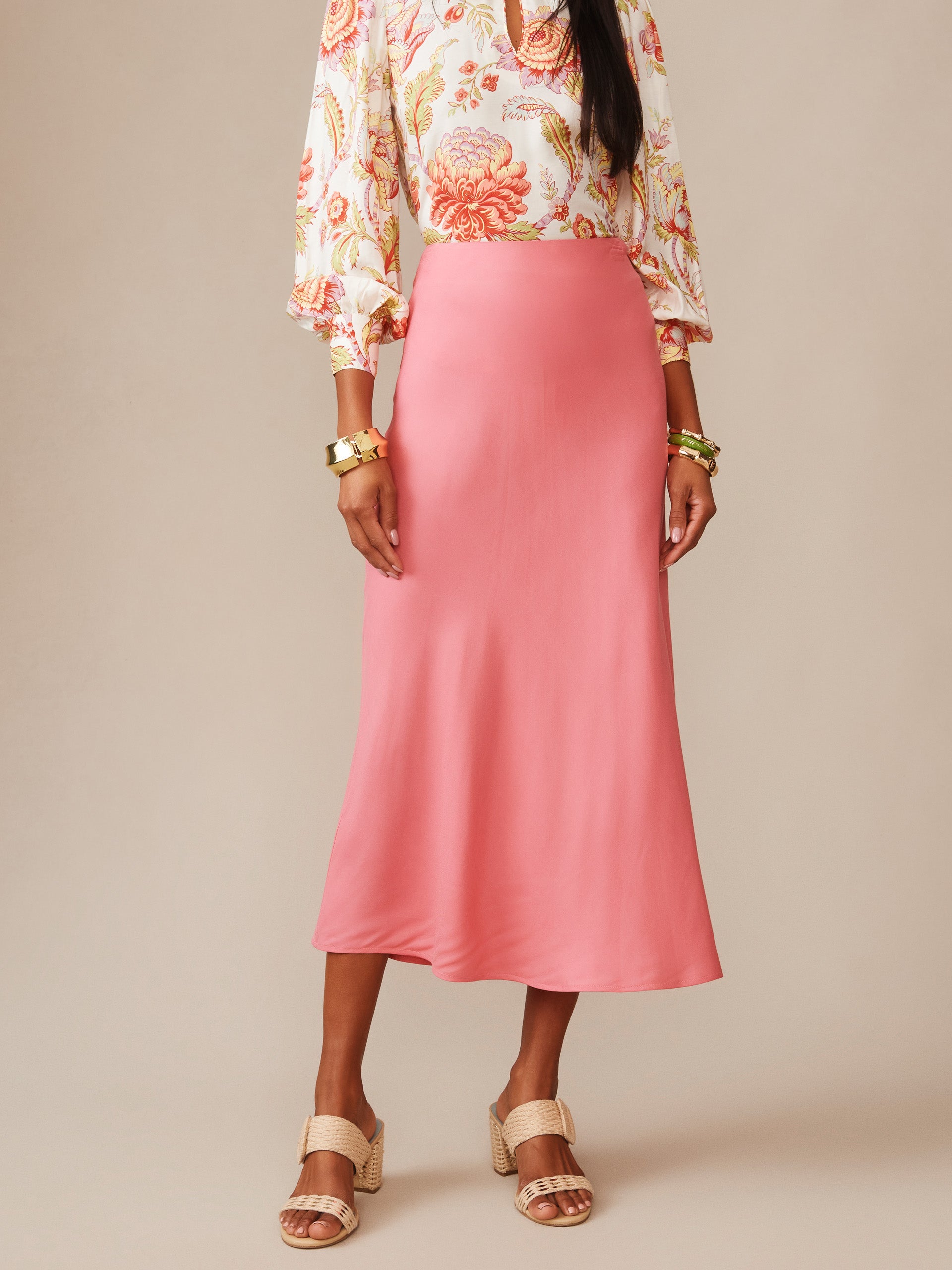 Model wearing a soft coral Adelyn Silk Blend Skirt by J.McLaughlin paired with a patterned blouse and woven sandals, showcasing effortlessly stylish, enduring designs.