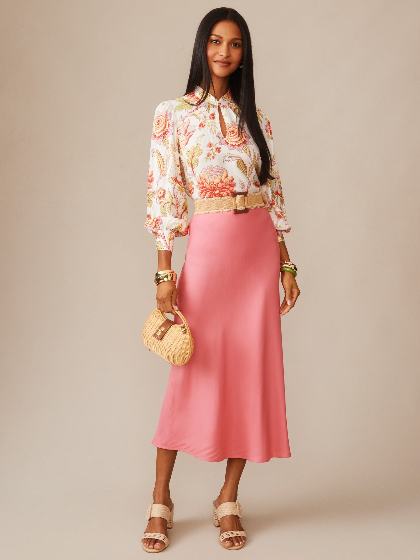 Woman wearing a coral pink Adelyn Silk Blend Skirt by J.McLaughlin paired with a floral blouse, beige sandals, and a woven handbag, showcasing effortlessly stylish, enduring designs.
