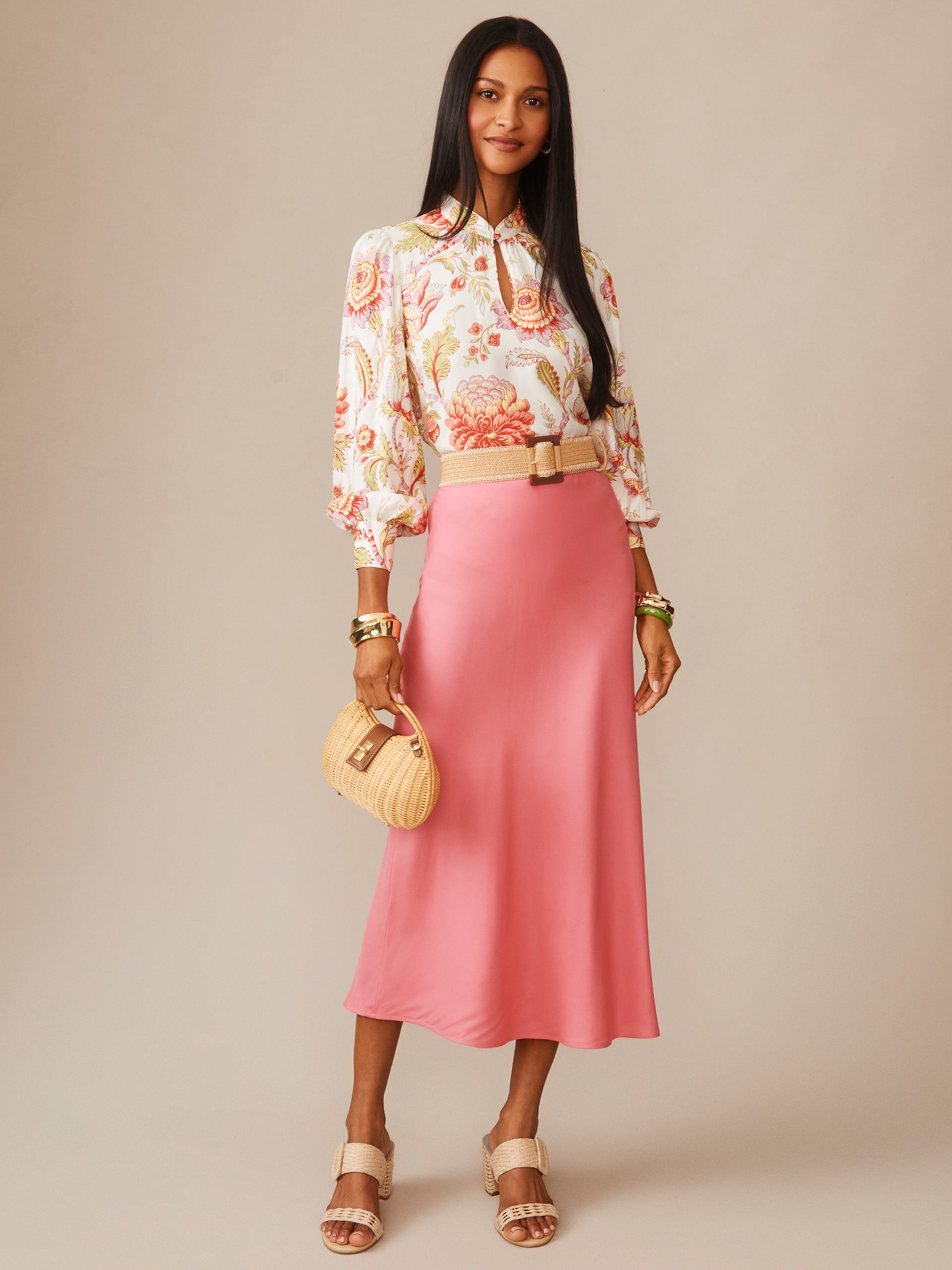 Woman wearing a coral pink Adelyn Silk Blend Skirt by J.McLaughlin paired with a floral blouse, beige sandals, and a woven handbag, showcasing effortlessly stylish, enduring designs.