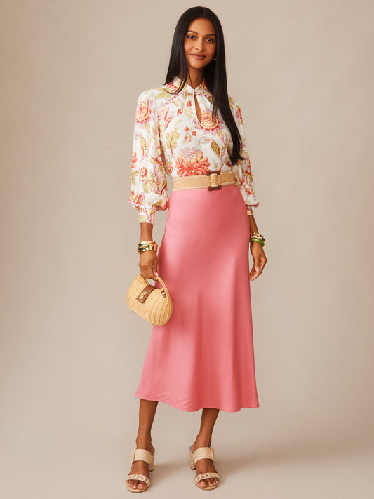 Woman wearing a coral pink Adelyn Silk Blend Skirt by J.McLaughlin paired with a floral blouse, beige sandals, and a woven handbag, showcasing effortlessly stylish, enduring designs.
