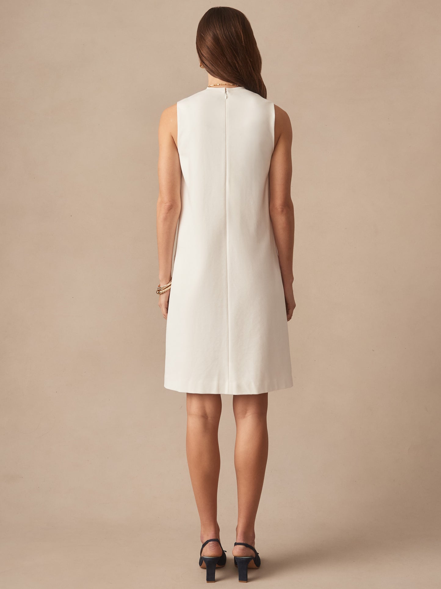 A woman stands facing away wearing a sleeveless ivory Alicia Dress by J.McLaughlin, showcasing effortlessly stylish, enduring designs with innovative fabrics and a legacy of timeless fashion.