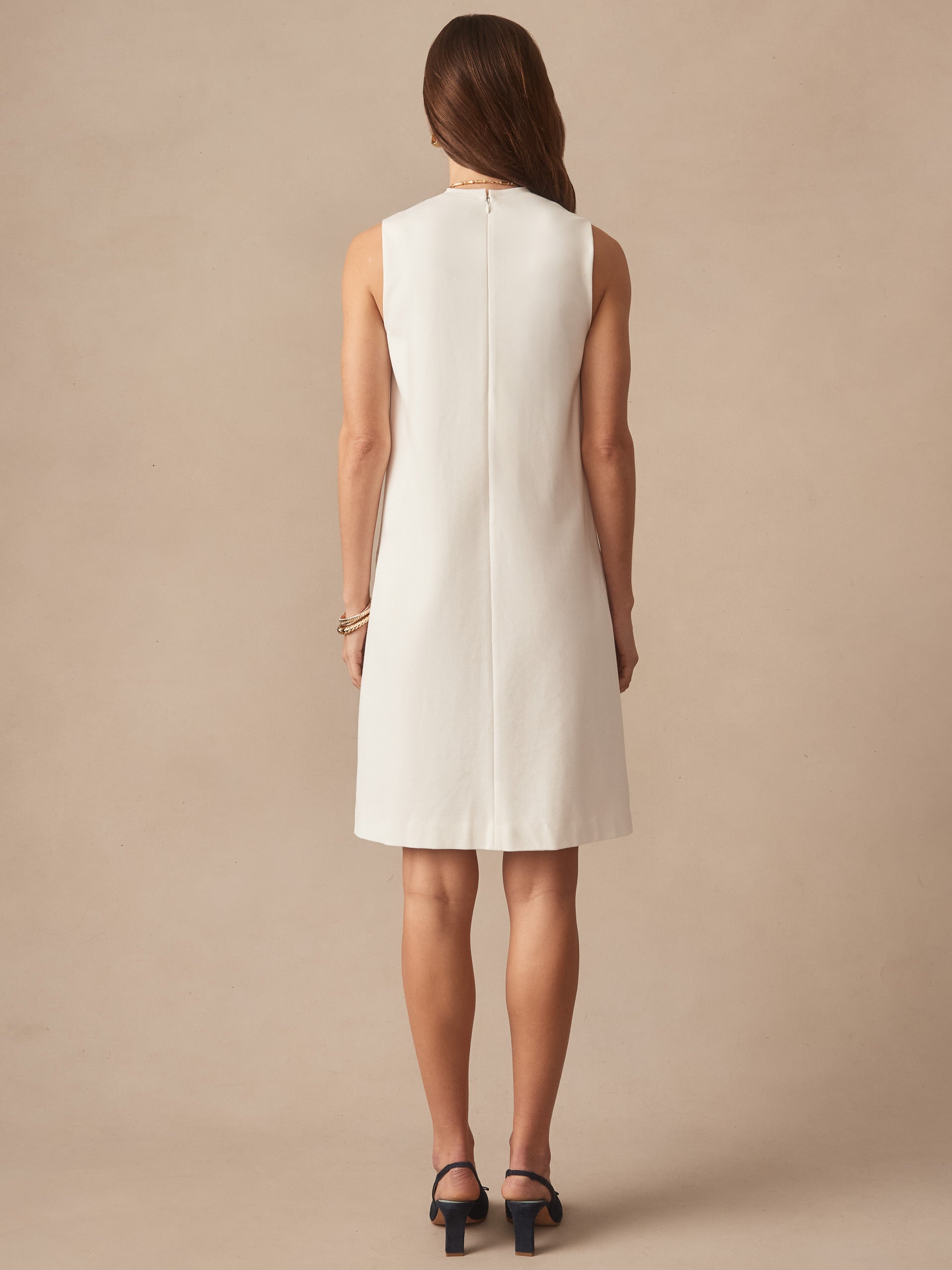 A woman stands facing away wearing a sleeveless ivory Alicia Dress by J.McLaughlin, showcasing effortlessly stylish, enduring designs with innovative fabrics and a legacy of timeless fashion.