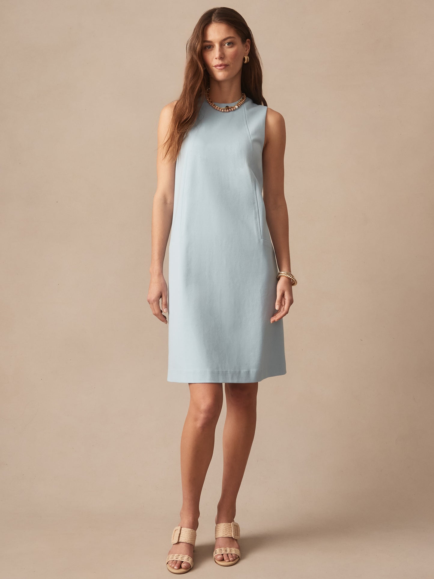 A woman wearing a sky blue Alicia Dress by J.McLaughlin, styled with gold jewelry and neutral sandals, showcasing effortlessly stylish, enduring designs and innovative fabrics.